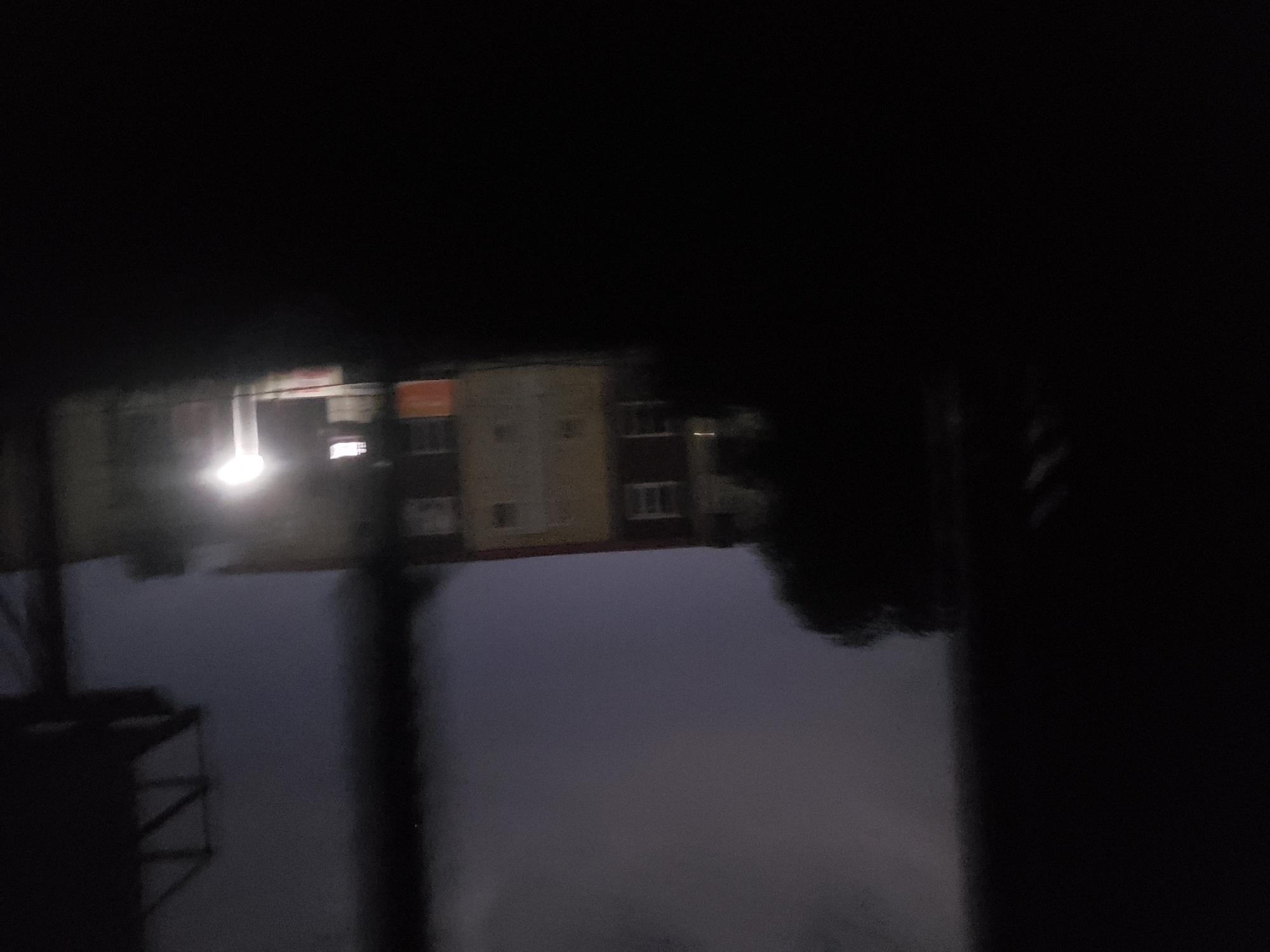 The image depicts a blurry, dimly lit urban scene at night. In the background, a multi-story building with visible windows and an orange-colored section can be seen. A bright light source illuminates a portion of the building and its surroundings. To the right of the building, a dark, angular structure resembling a billboard support is visible against the dusky sky. In the foreground and to the left, large, dark, out-of-focus shapes obscure the view, suggesting the presence of an object or structure closer to the camera. The overall impression is one of a nighttime street view in an urban area, with limited visibility due to darkness and motion blur. The location is identified as Iganmu, Nigeria.