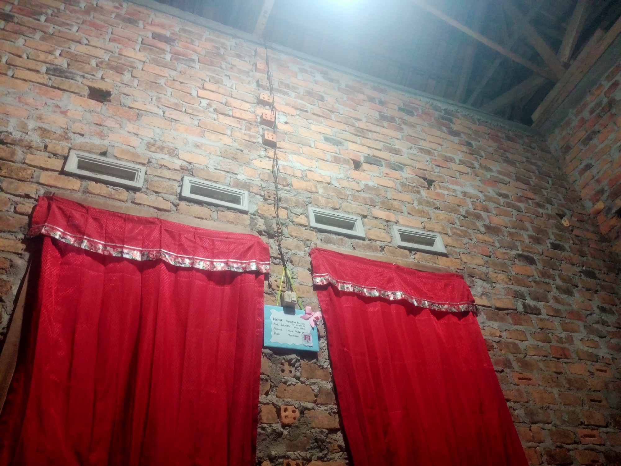 The image depicts an interior view of a room with a brick wall. Two red curtains with decorative trim are hung on the wall, flanking a small framed sign. The sign, which appears to be a personal information board, has handwritten text in Indonesian, including fields for "Nama" (Name), "Asal Sekolah" (School Origin), "Alamat" (Address), "Hobi" (Hobby), and "Impian" (Dream). There are also small windows or vents visible in the brickwork above the curtains. The ceiling above shows exposed wooden beams. The lighting suggests it is nighttime or a dimly lit indoor space. The scene is likely within a residential setting in Nusa Agung, Indonesia, given the provided context.