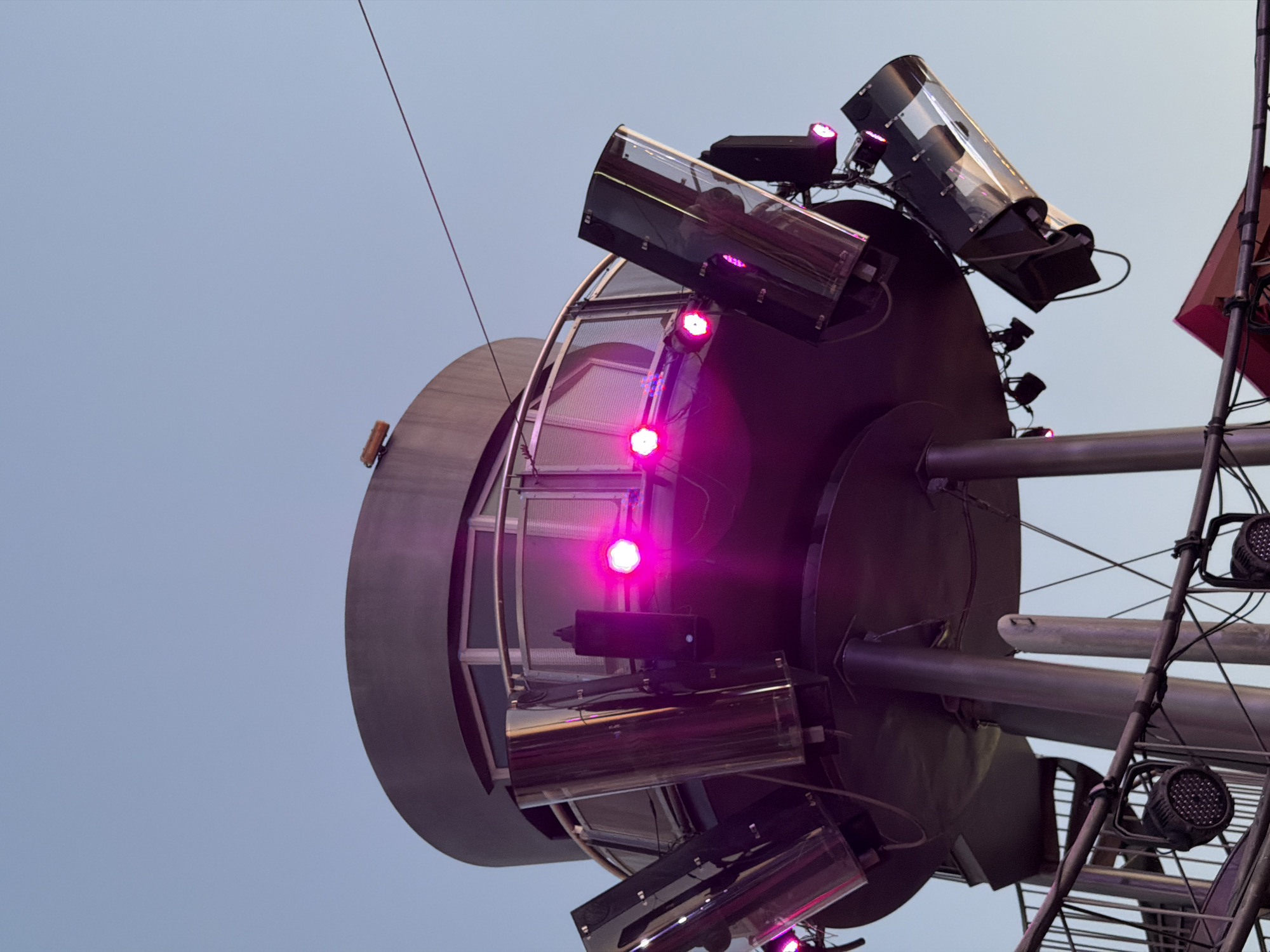 The image captures a low-angle view of a futuristic-looking structure, possibly a concert venue or observation deck, at night in Las Rozas de Madrid, Spain. The structure features a circular, multi-tiered design with metallic columns supporting an upper level that has a glass-enclosed observation area. Several stage lights, some emitting bright pink illumination, are mounted around the exterior of the upper level and below it, suggesting an event is taking place or has recently concluded. A thin wire stretches across the sky above the structure. The sky is clear and a pale blue, indicating twilight or a very early morning. The overall impression is one of modern architecture and entertainment infrastructure.