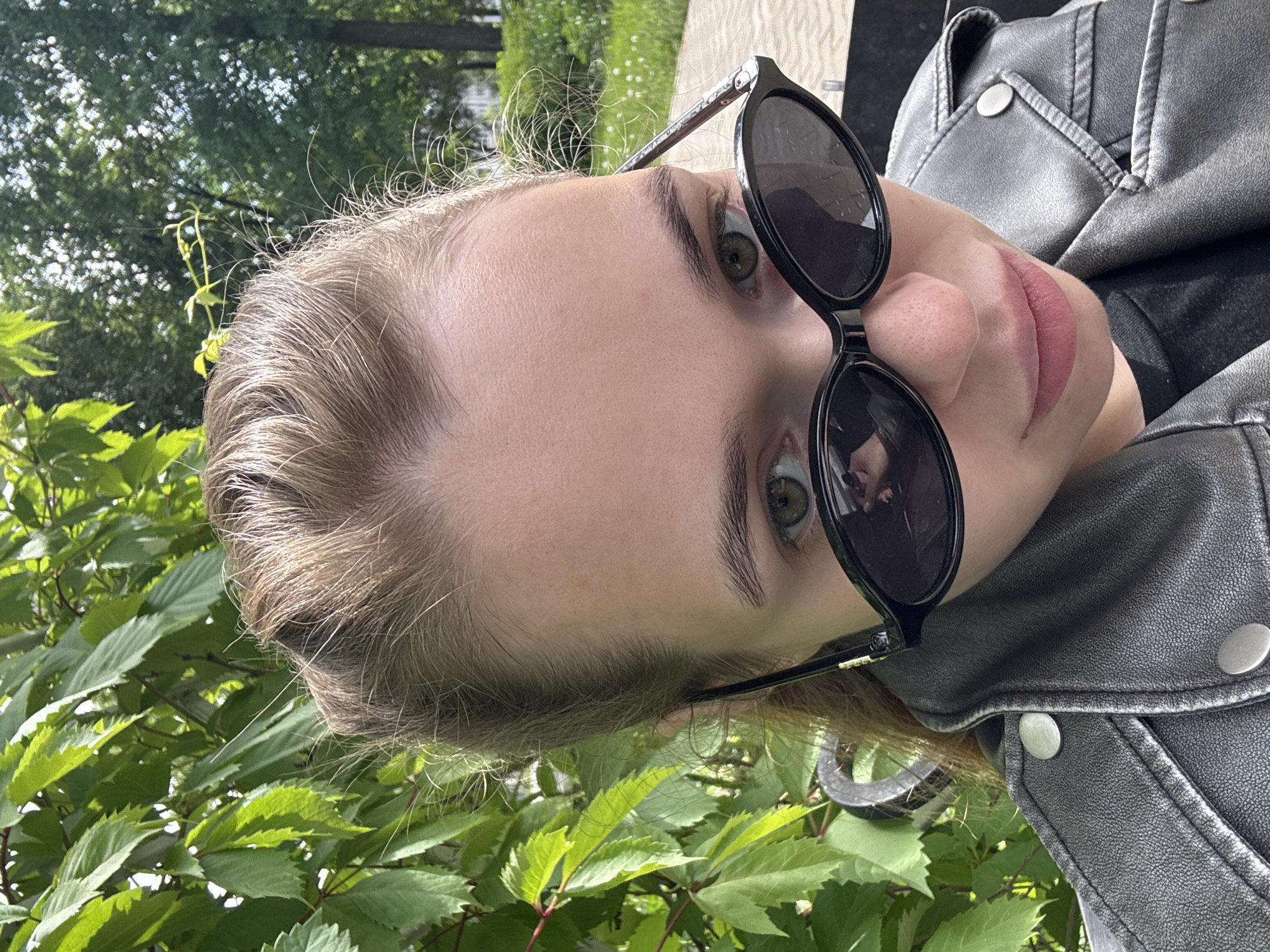A person with light skin and blonde hair is shown wearing sunglasses and a gray leather jacket. The person's face is tilted slightly upwards towards the camera, with green foliage visible in the background. The scene appears to be outdoors, possibly in a park or garden setting, given the presence of green leaves and vegetation. Based on the presence of green foliage, it may be spring or summer. There is nothing in the image to determine if the location is in Chaikovsky, Russia.