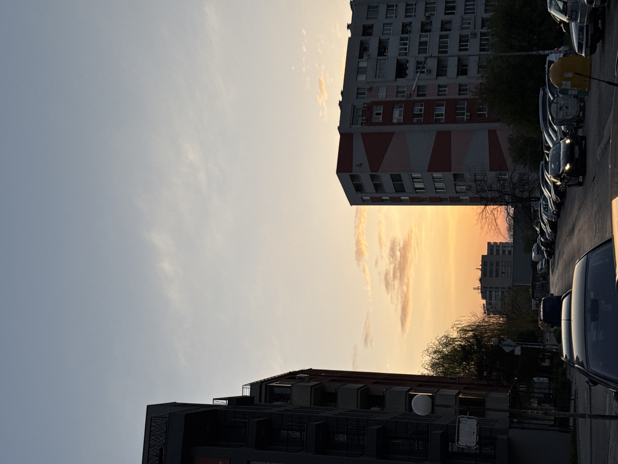 This is an outdoor, medium shot taken in Sofia, Bulgaria. The image shows two buildings, one modern and the other a Soviet-era apartment block, during a sunset. The modern building is situated in the foreground, while the Soviet-era building is in the background. A row of parked cars is visible on the right. The sky shows a clear transition between the light blue day sky to the left and the orange sunset clouds at the center. The scene is quiet with no visible people or active interactions.