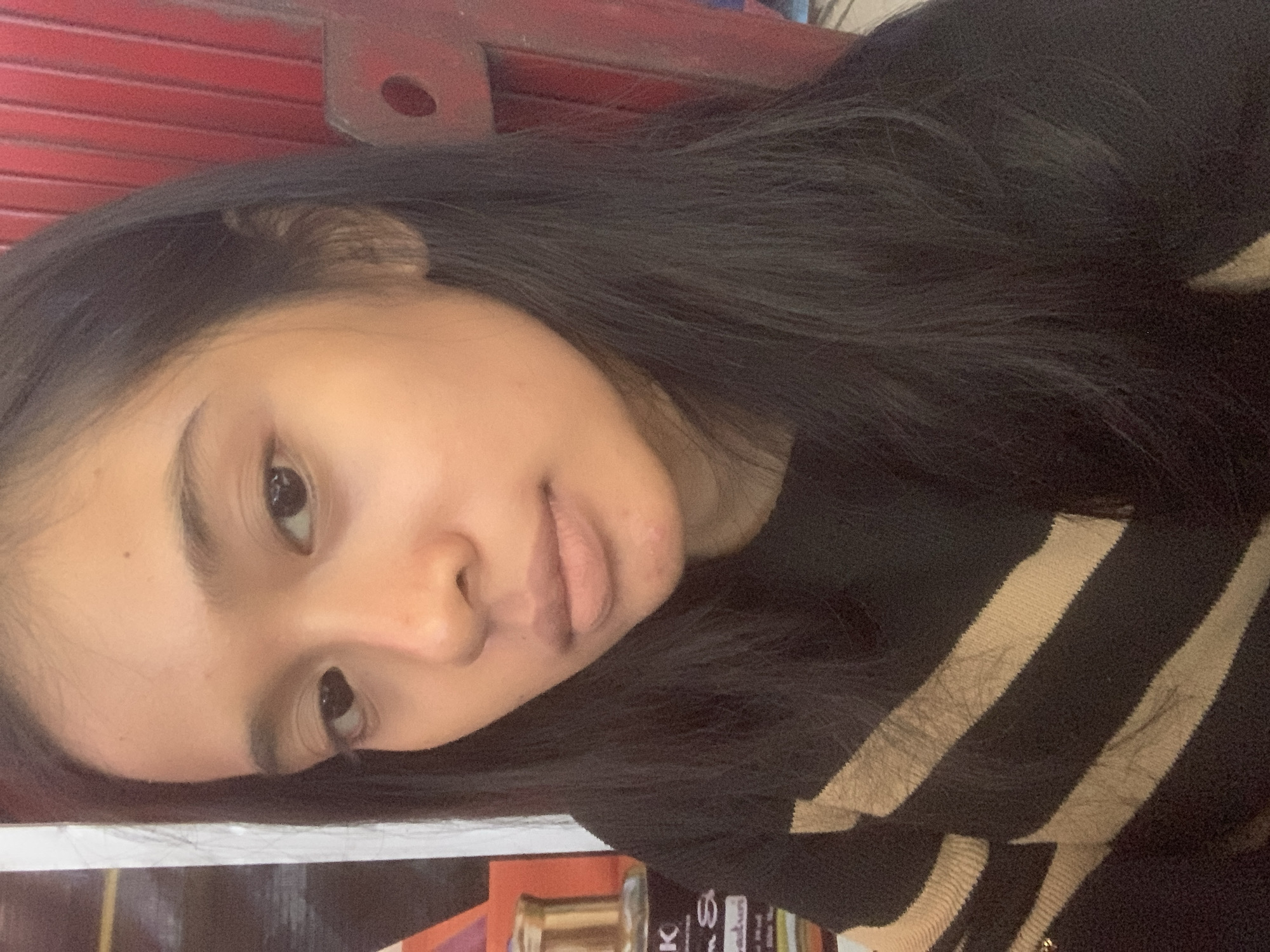 A portrait-style photograph, originally rotated, features a young individual, likely female, captured from the chest up. She has long, dark, straight hair, dark eyes, and a neutral to slightly gentle expression with full lips. Her head is subtly tilted, and she gazes directly at the viewer. She is dressed in a black top adorned with broad horizontal beige or light brown stripes.
The blurred background behind her head reveals a textured, reddish surface, possibly a wall or shutter, with a dark, circular recess. Below her, out of focus, are indications of objects on a surface, likely a shelf or counter. A golden-colored bottle or container is partially visible, alongside fragmentary text that includes the letters "K" and "G S". The lighting is soft and appears to be internal or diffused. No specific activity is shown beyond posing for the image. The location context is Sungai Meriam, Indonesia.