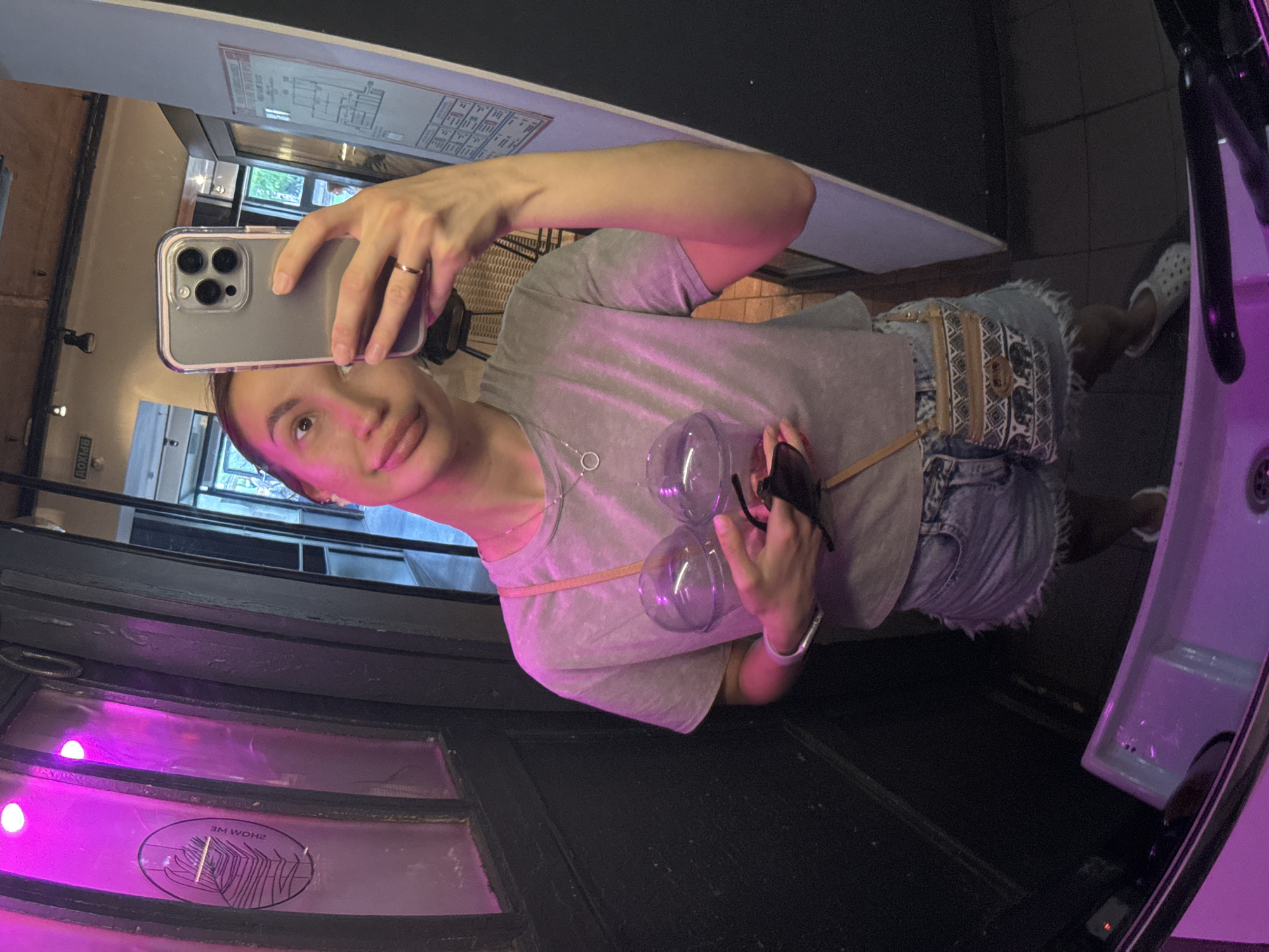 A person is taking a selfie in a mirror. The individual, presumably female, is holding a grey smartphone up with the left hand. The person is wearing a grey shirt, jean shorts, and a bracelet. A purse is slung over the shoulder. The reflection in the mirror shows part of the room's interior, which has a window with a display featuring neon lights. A plan is visible in the upper portion of the mirror. The background includes a sink, indicating the setting is likely a restroom. According to text on the window, the location is in Nizhny Novgorod, Russia.