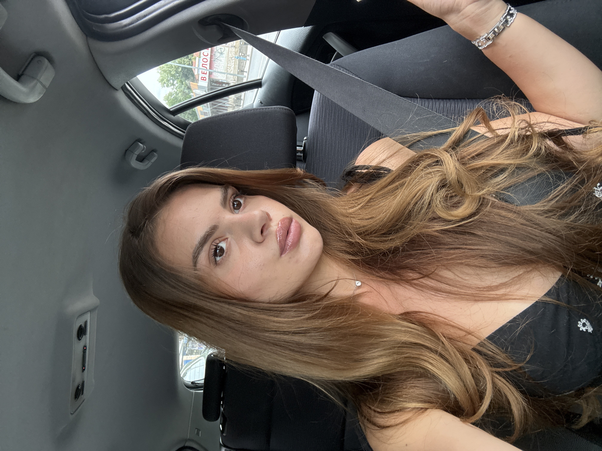 A young woman with long, wavy brown hair is sitting in the passenger seat of a car. She is wearing a black top and a silver necklace with a small pendant. A seatbelt is visible across her chest. Outside the car window, there are trees and buildings. One building has a red and white sign with Cyrillic text that reads "ВЕЛОС" (Velos). This sign is on the facade of a building located at Hristo Botev Blvd. 17 in Plovdiv, Bulgaria, which houses a bicycle repair shop and showroom. The image is taken from a selfie perspective, suggesting the woman is taking the photograph herself.