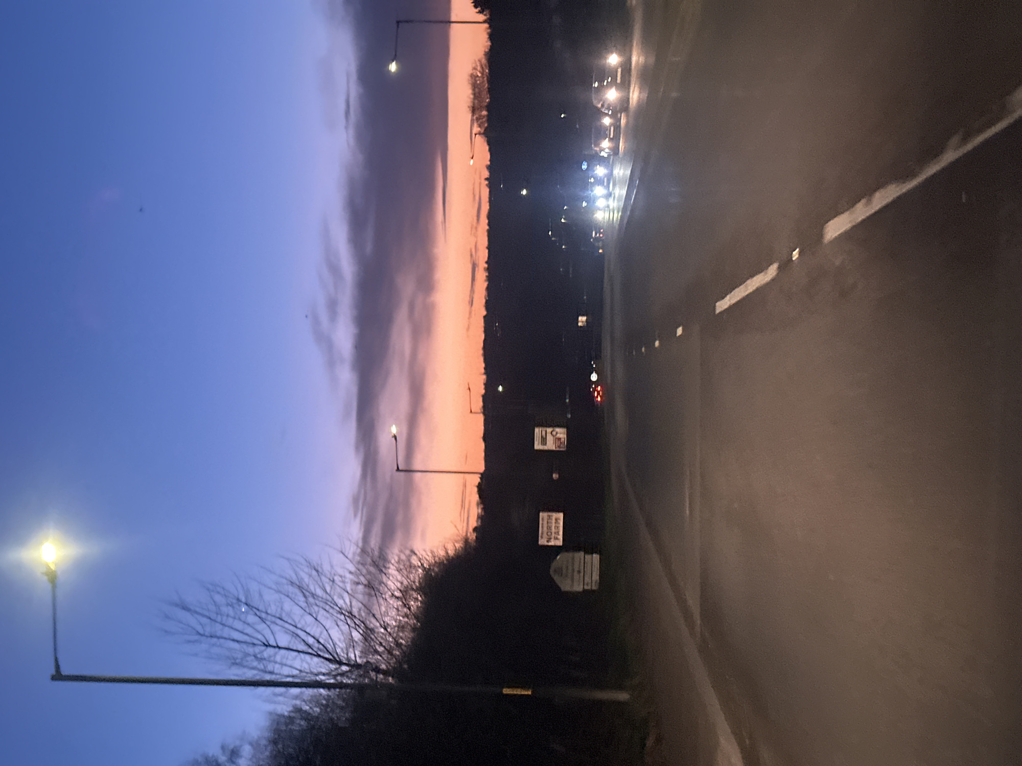 The image depicts a twilight scene on a road in Tunbridge Wells, United Kingdom. The sky transitions from deep blue overhead to an orange and red horizon, partially obscured by dark clouds. The asphalt road, which appears damp in areas, has two visible lanes separated by dashed white lines, extending into the distance. Numerous vehicles with illuminated headlights are traveling towards the viewer in the opposing lane, creating significant glare. On the left side of the road, a bright streetlight is illuminated, alongside bare deciduous trees and dense dark foliage. A white sign with "NORTH FARM" in dark lettering is visible on the left verge, accompanied by additional indistinct signage. Another streetlight is visible further down the road on the left, and several more line the right side of the road in the distance. No individuals are visible.