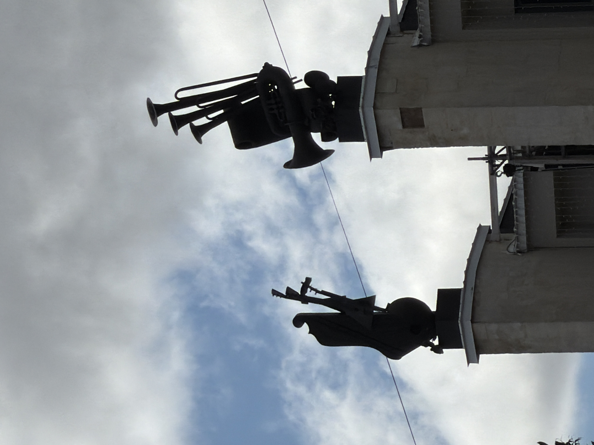 A low-angle shot shows two trumpet sculptures on buildings in Izhevsk, Russia, against a cloudy sky. The sculptures are dark silhouettes. The buildings are light grey concrete structures, with windows visible on the right side of the frame. A thin wire runs diagonally across the sky between the sculptures.