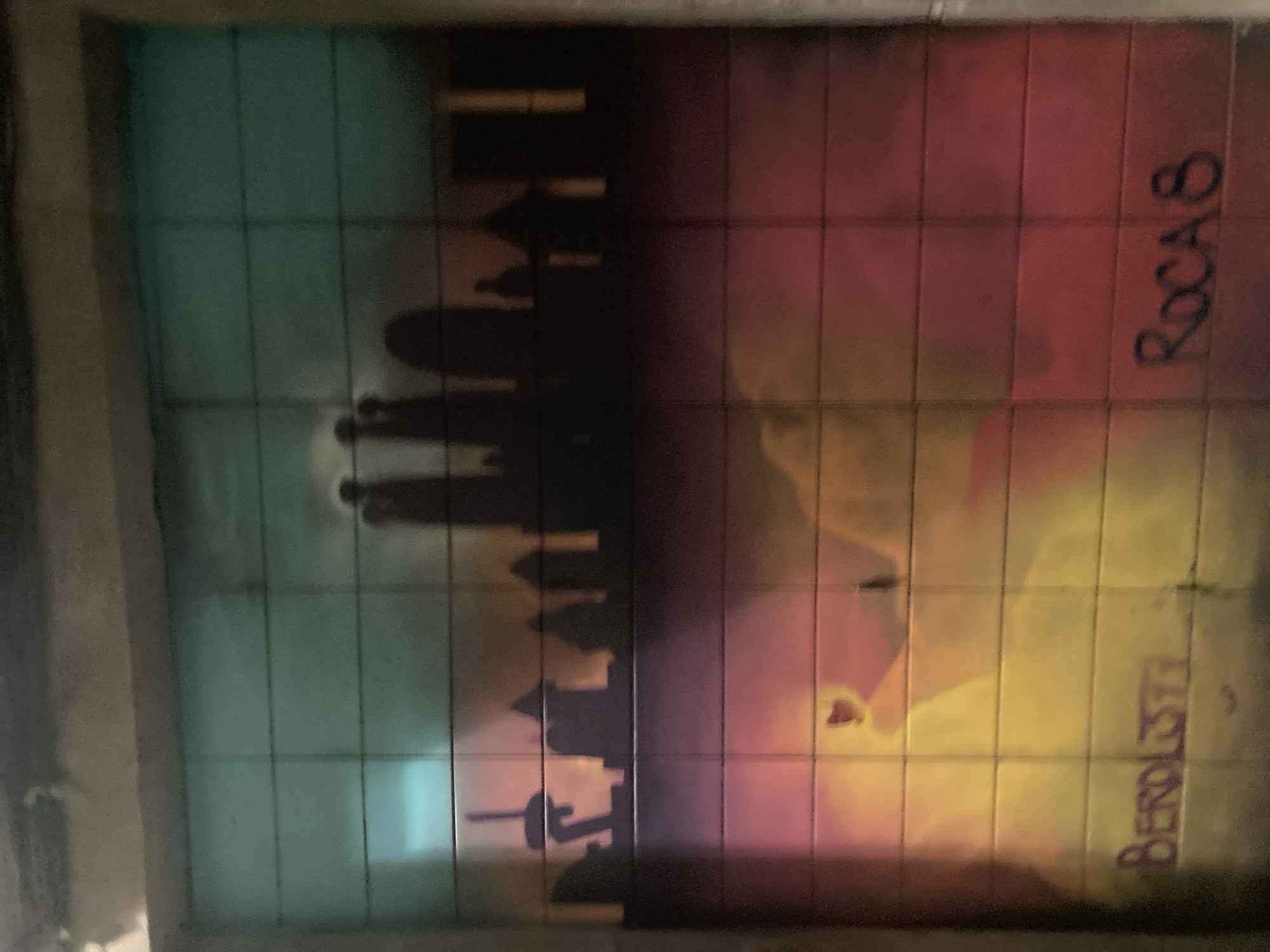The image is a photograph of a graffiti mural on a tiled wall, depicting a silhouetted skyline of buildings against a sunset or sunrise sky. The sky transitions from a teal and green hue at the top to shades of pink, orange, and yellow towards the bottom. The skyline includes various structures with pointed roofs and at least one prominent tower resembling a church spire. In the lower portion of the mural, graffiti tags are visible. On the left, the tag "BEROLDSIF" is written in purple, and on the right, "ROCA 8" is written in black. The tiled surface of the wall is evident, with visible grout lines. The overall scene suggests an urban environment, likely in Barcelona, Spain, given the architectural style hinted at in the graffiti.