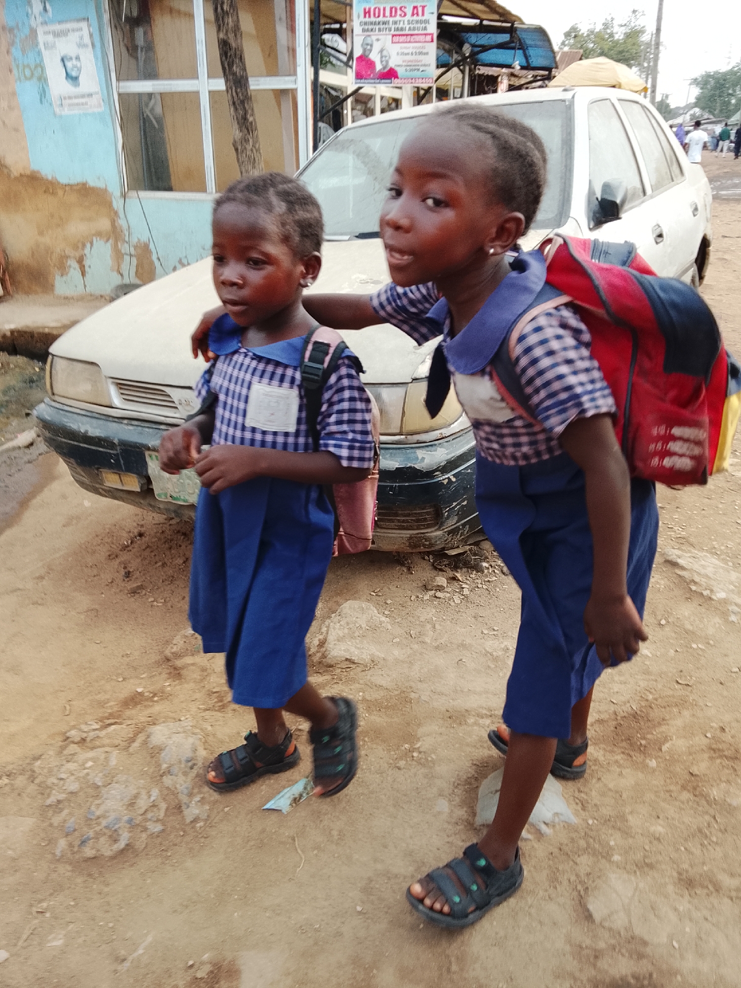 Two young, Black schoolgirls in matching blue and plaid school uniforms and dark sandals walk on a dirt road. One girl has her arm around the other's shoulder. They both wear backpacks. A white car is parked behind them. In the background are buildings and people. A sign on a building reads, "HOLDS AT - CHINWOKE INT'L SCHOOL DAKI BIYU JABI AREA ABUJA." The location is in the Daki Biyu Jabi area of Abuja, Nigeria.