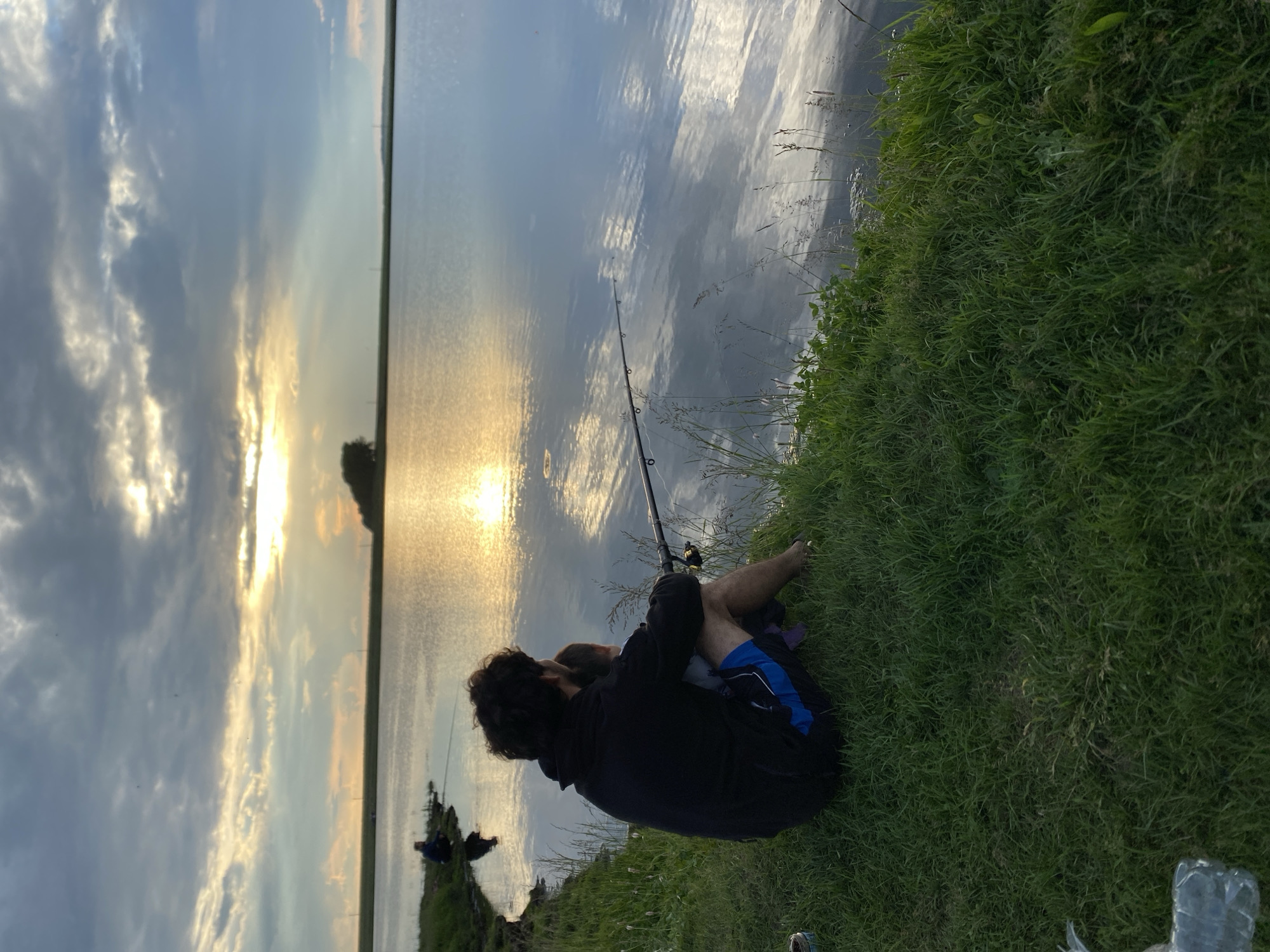 In the foreground, a person with dark, curly hair sits on lush green grass, facing a tranquil body of water. They are wearing a black hooded sweatshirt and blue and black shorts. The person is holding a fishing rod, with the line extending into the water. A child, partially visible, is seated in front of them, also facing the water. The sun is setting, casting a golden glow on the water and painting the sky with dramatic clouds. In the middle ground, other figures can be seen fishing along the shoreline in the distance. The environment appears to be a natural landscape, possibly a lake or reservoir, on the outskirts of Сыйрышбашево, Russia. The overall atmosphere is peaceful and serene.