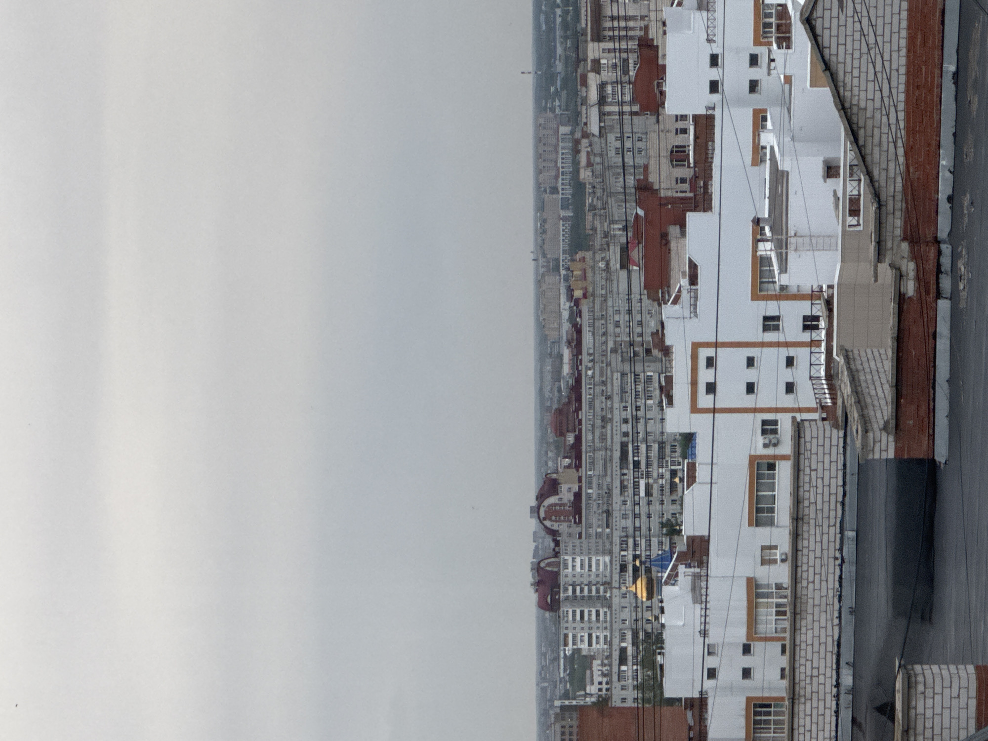 The image shows a cityscape, likely taken from a high vantage point such as a tall building. The sky is overcast. In the foreground, there is a building with white walls and brown trim along the windows and edges. The building is made of brick. Power lines are visible. The buildings in the distance are gray and have multiple stories. A structure with a gold dome is visible in the distance.