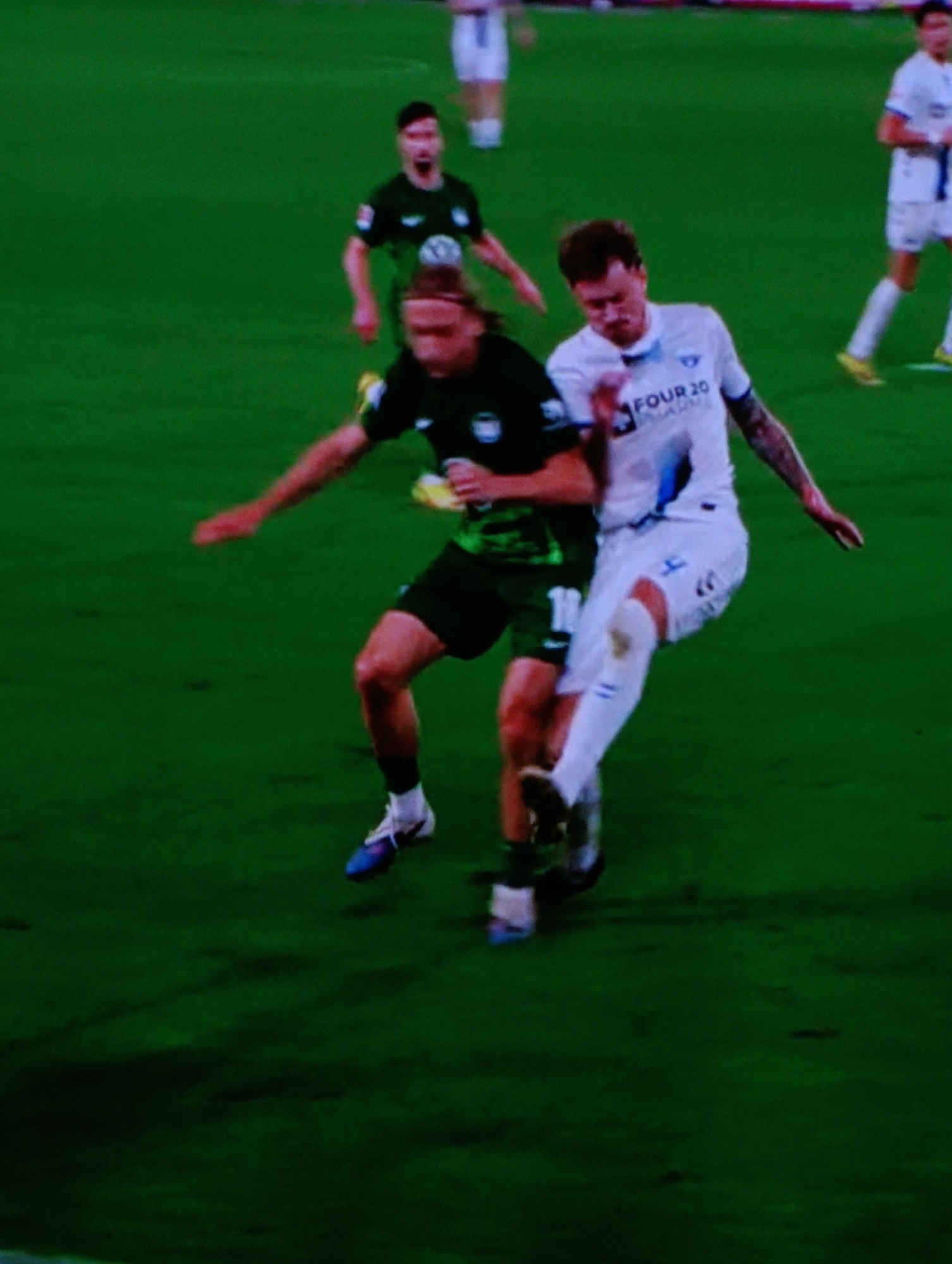 On a green sports field, two soccer players vie for the ball. The player on the left wears a dark green jersey and shorts with blue and white cleats. He extends his left arm outward. The player on the right is in a white jersey and shorts with white socks and dark cleats. His right leg is extended towards the ball. Other players are visible in the background. The location is the city of Plovdiv, Bulgaria.