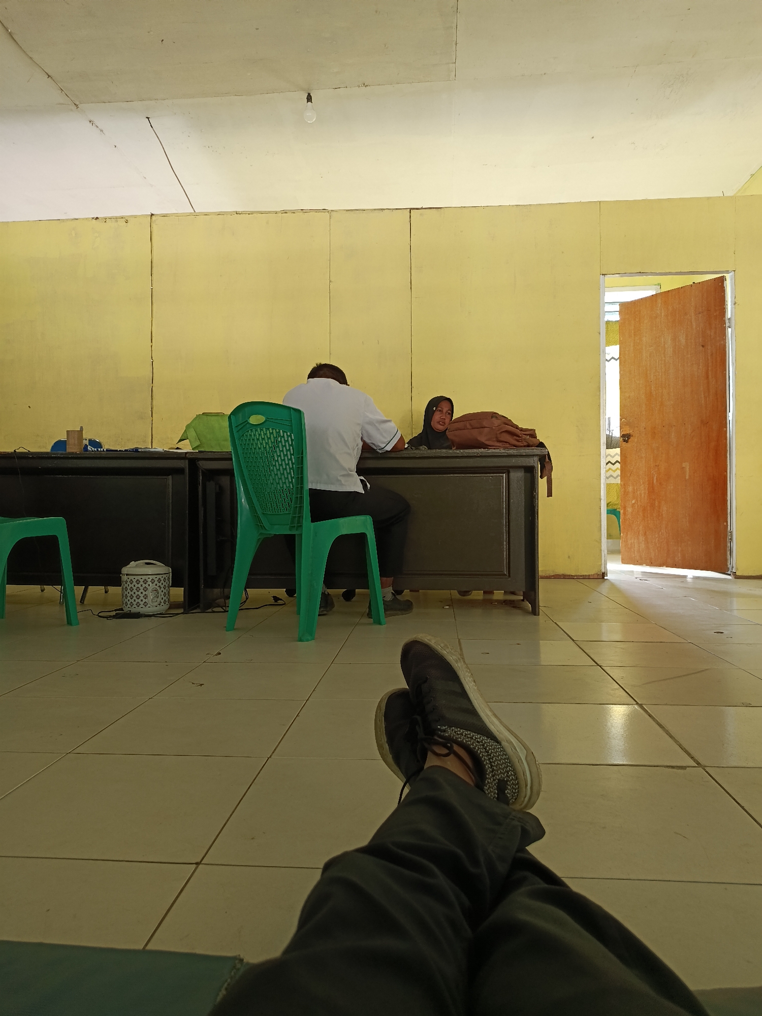 The image depicts an indoor scene, possibly an office or classroom, in Mansalean, Indonesia. The walls are painted a muted yellow, and the ceiling is white with exposed wires and a single light bulb. The floor is tiled with large, light-colored squares. In the foreground, a person's legs are visible, crossed and wearing dark pants and athletic shoes.
The mid-ground of the image features two desks arranged parallel to each other, facing the viewer. On the left desk, a white electric rice cooker is visible. A green plastic chair is positioned between the desks, with its back to the camera. A man wearing a white shirt and dark pants is seated at the right desk, facing away from the viewer. He appears to be looking down at something on the desk. To his right, a woman wearing a black headscarf and a black top is seated at the same desk. She is looking towards the camera with a neutral expression. A brown backpack rests on the desk next to her. Another green plastic chair is visible to the far left of the frame, in front of the left desk.
To the right of the desks, a wooden door is ajar, revealing a glimpse of a brightly lit space beyond, possibly another room or hallway. The light filtering through the doorway casts shadows on the floor. The overall atmosphere appears to be casual and somewhat relaxed, given the posture of the person in the foreground and the open door.