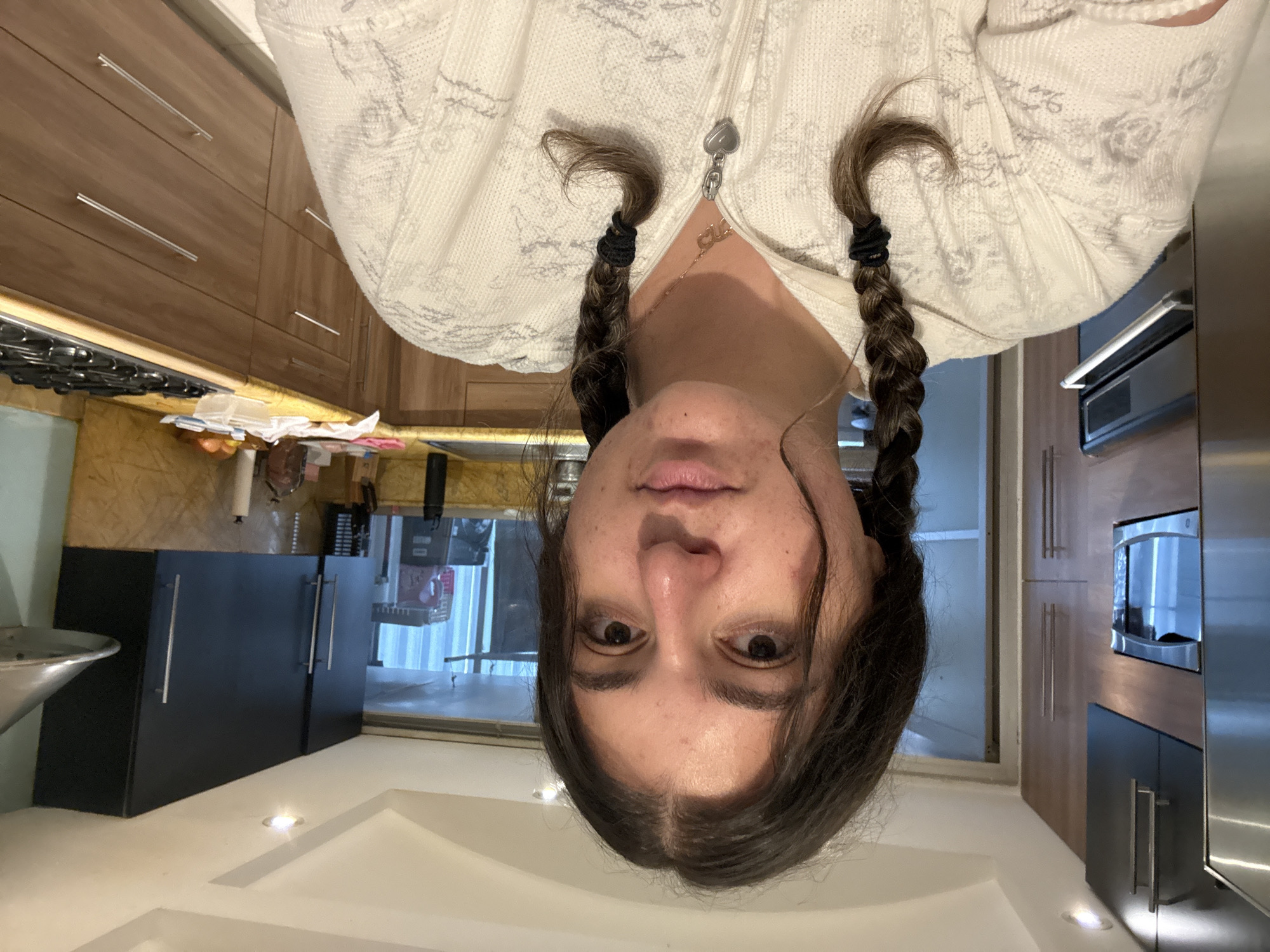 The image shows a woman with brown hair braided into two pigtails, wearing a white patterned zip-up sweater, facing the camera. She appears to be inside a kitchen with wooden cabinets and black lower cabinets. A stainless steel appliance (likely a refrigerator or oven) is visible on the right side of the image. The location is likely Mexico City, Mexico.