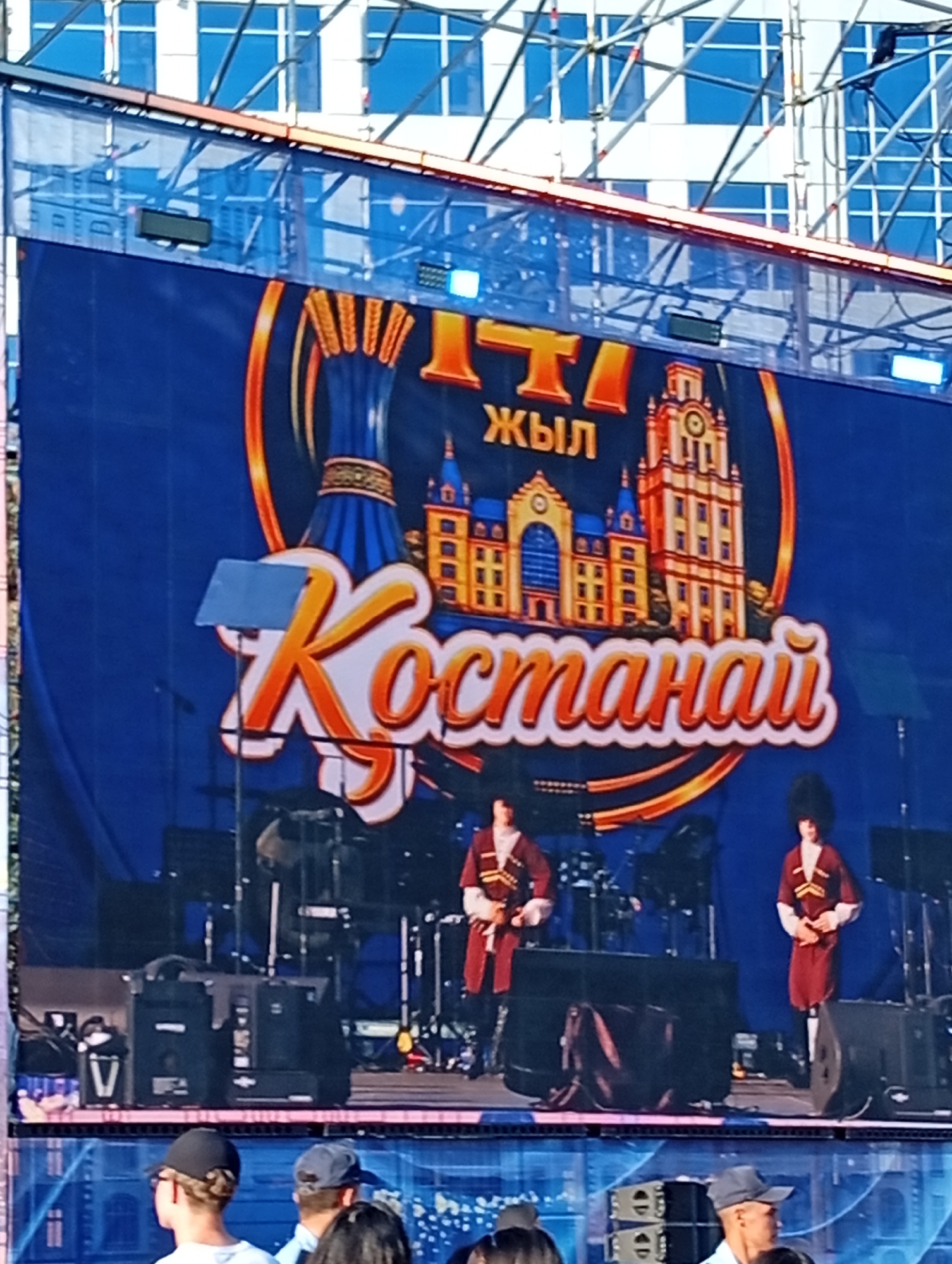 This is a medium-shot, slightly blurry, daytime outdoor image of a festival or performance stage in Kostanay, Kazakhstan. The dominant element is a large blue banner with a festive golden graphic. In the center of the banner is the word "Kostanay" written in large, stylized orange and white letters, with a golden outline. Above this, in smaller orange letters, are the Cyrillic characters "ТЫ ЖЫЛ" which translate to "This Year." Flanking the text are illustrations of ornate buildings, reminiscent of historic architecture, and a depiction of a traditional Kazak yurt or decorative element.
On the stage, visible in the lower half of the image, two performers are dressed in traditional Kazak attire. They wear red robes with decorative trim and tall, furry hats. The performer on the left is holding a long object, possibly a staff or musical instrument, and is looking towards the right. The performer on the right is standing with their hands clasped in front of them, looking forward. Behind the performers, drum kits and other musical equipment are set up. In the foreground, at the bottom of the image, several audience members with hats are visible from the back, looking towards the stage. The backdrop behind the stage appears to be a modern building with a grid of windows. The overall impression is a lively cultural event.