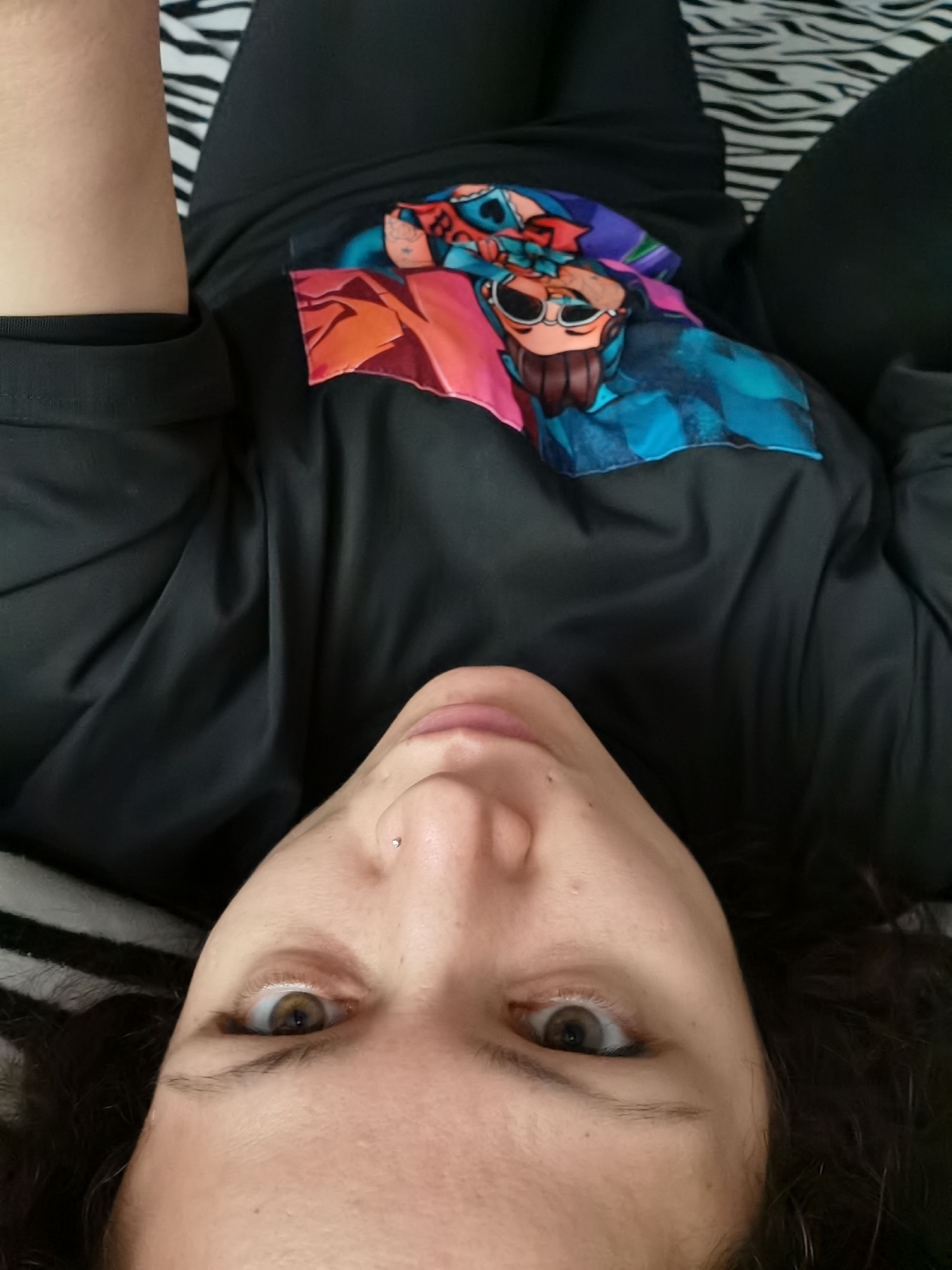 An upside-down, close-up, eye-level shot focuses on the face of a woman lying on a striped surface. The woman has brown eyes, dark hair, and a nose piercing. She is wearing a black t-shirt with a colorful graphic of a cartoon character. The background consists of black and white stripes, possibly a bedding or upholstery. The image does not contain information that would allow for its localization within the city of Chaikovsky, Russia.