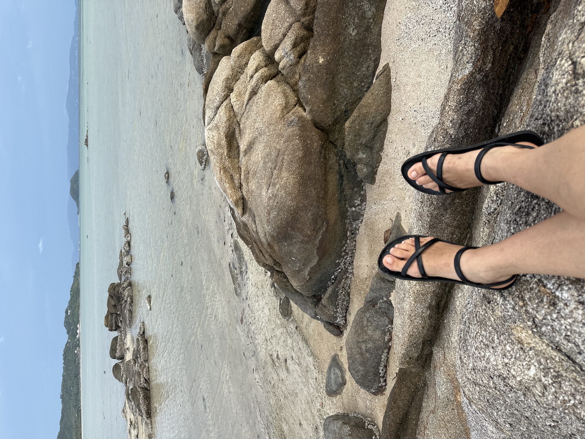 The image captures a pair of bare legs and feet adorned with black strappy sandals, resting on a textured rock. The setting appears to be a beach in ตำบลเกาะพะงัน, Thailand, as evidenced by the presence of rocks, sand, and the calm sea in the background. The sky is overcast, adding to the somewhat muted color palette of the scene. The focus is on the footwear and the textures of the surrounding natural elements.