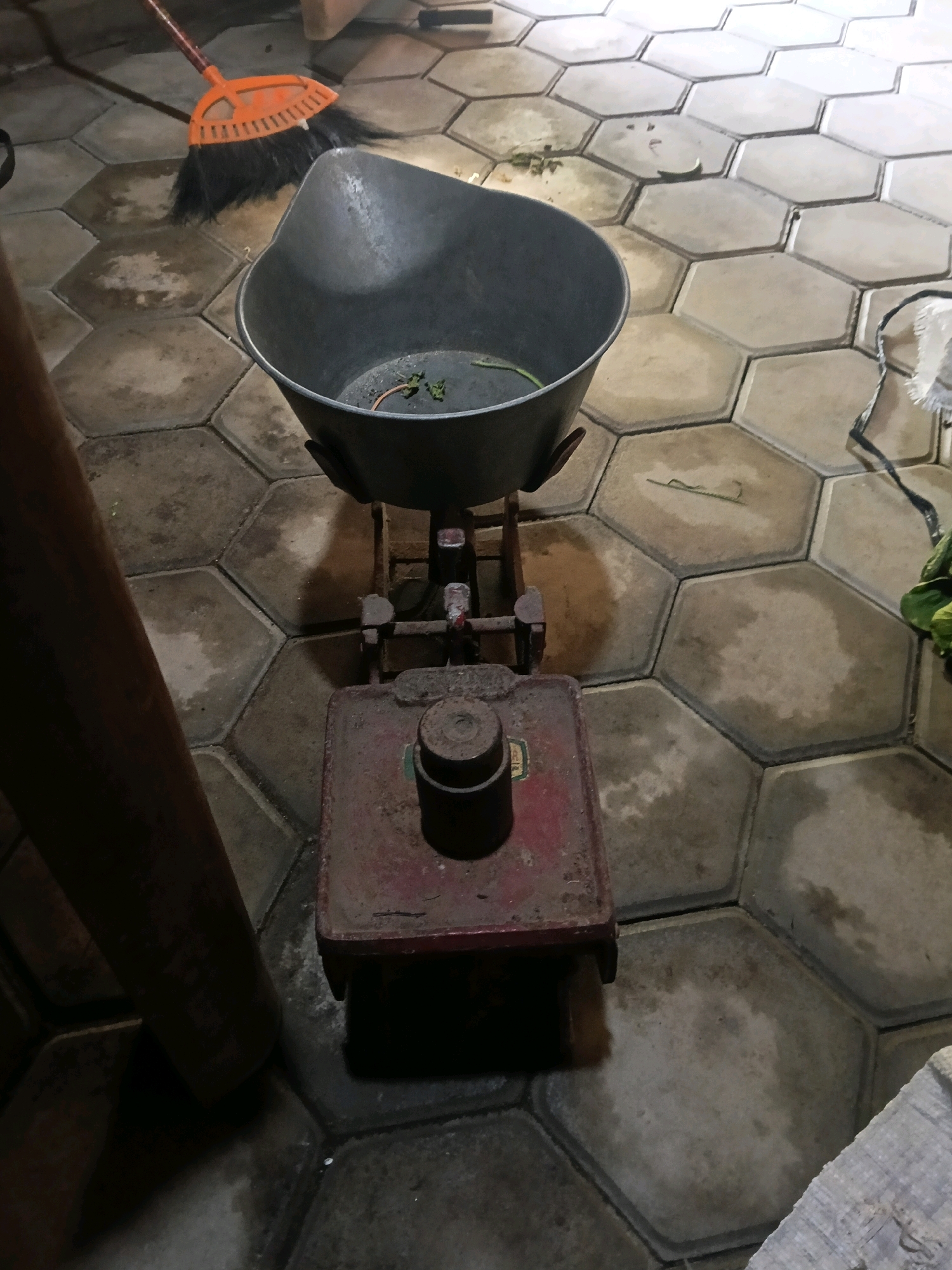 The image depicts an outdoor scene on a tiled ground, possibly in Nglungger, Indonesia. In the foreground, a vintage industrial scale is prominently featured. The scale's base is made of red metal, showing signs of rust and wear. A cylindrical weight, also metal, rests on the scale's platform. Above the base, a metal beam structure supports a large, dark gray metal basin. The basin appears to be empty except for some dry leaves and small debris scattered at its bottom.
To the left of the scale, a large, dark brown, cylindrical object stands vertically, partially obscuring the background. Behind the scale and to the left, an orange broom with black bristles lies on the ground, its handle pointing upwards and to the left. The ground is covered with hexagonal concrete tiles, some of which are wet or discolored, suggesting recent cleaning or exposure to moisture. The lighting in the scene is dim, creating shadows and highlighting the textures of the objects. The overall impression is one of a utilitarian space, perhaps a backyard or an outdoor storage area.