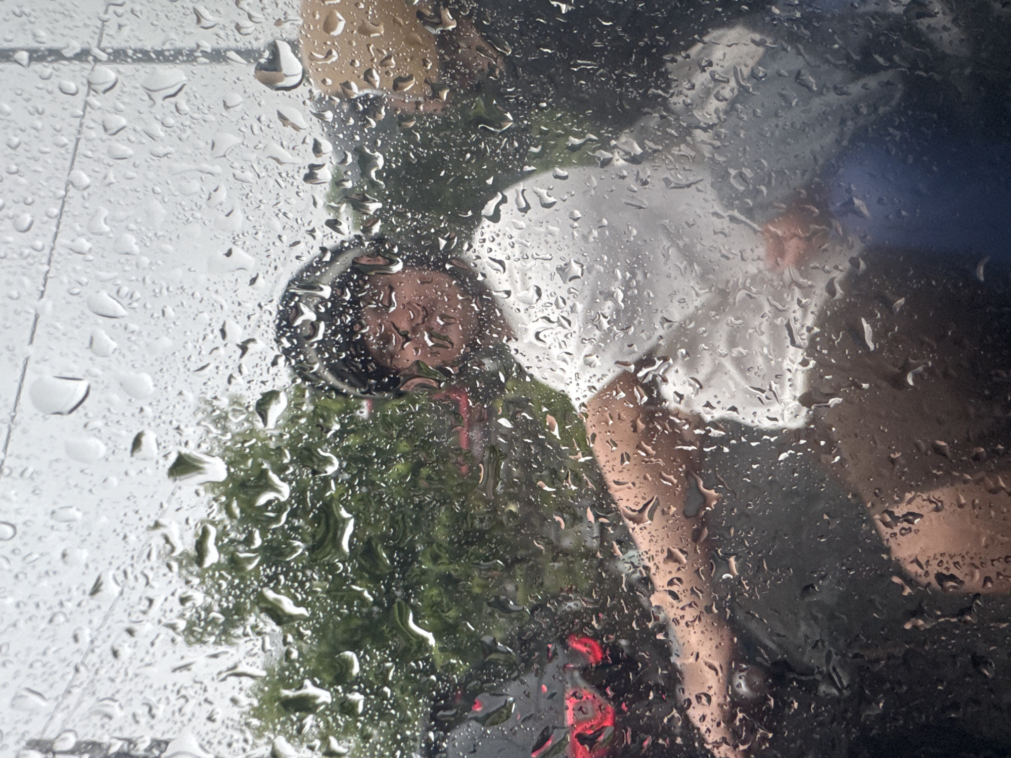 The image presents a view through a wet glass surface, likely a window or windshield, covered extensively with numerous raindrops that distort the scene beyond. The perspective is from an interior looking outwards into a rainy environment in Kuta, Indonesia.
In the midground, at least two human figures are partially visible. On the left, a person's head and upper body are discernible, appearing to wear a dark helmet or head covering, possibly on a motorbike. Their face is indistinct due to the water and angle. Their clothing seems to be dark green or black. To the right, another person is partially visible, clothed in a lighter, possibly white, garment, with an arm or hand showing a lighter skin tone.
The background features lush green foliage, suggesting trees or bushes, especially prominent in the center and left. Portions of what appears to be a light-colored paved ground or wall are visible in the upper left. A distinct bright red light, resembling a vehicle's taillight or brake light, glows near the bottom center, adding to the impression of a street scene. Other dark, indistinct shapes suggest the presence of additional vehicles or structures further away.
The activity taking place is difficult to pinpoint precisely due to the obscured view, but it appears to be people outdoors in an urban or semi-urban setting during rainfall. The weather is clearly rainy, indicated by the abundant water droplets. The time of day appears to be daytime, but overcast conditions are suggested by the diffused light. No legible text is visible in the image.