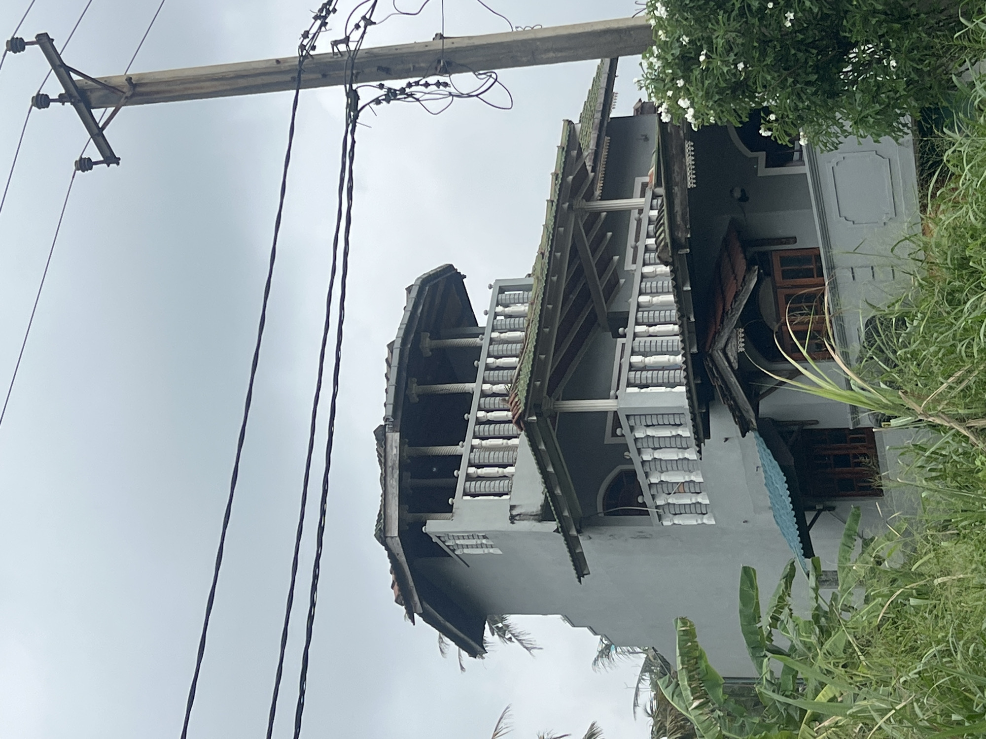 The image shows a two-story building located in Goviyapana, Sri Lanka. The building is painted gray with decorative white trim along the roof and balcony edges. The roof is multi-tiered and appears to be made of dark-colored tiles. A wooden utility pole with power lines runs along the left side of the image, partially obscuring the view of the building. Vegetation, including tall grasses and banana trees, is visible on the right side of the image, partially covering the building's ground level. The sky is overcast, suggesting a cloudy day.