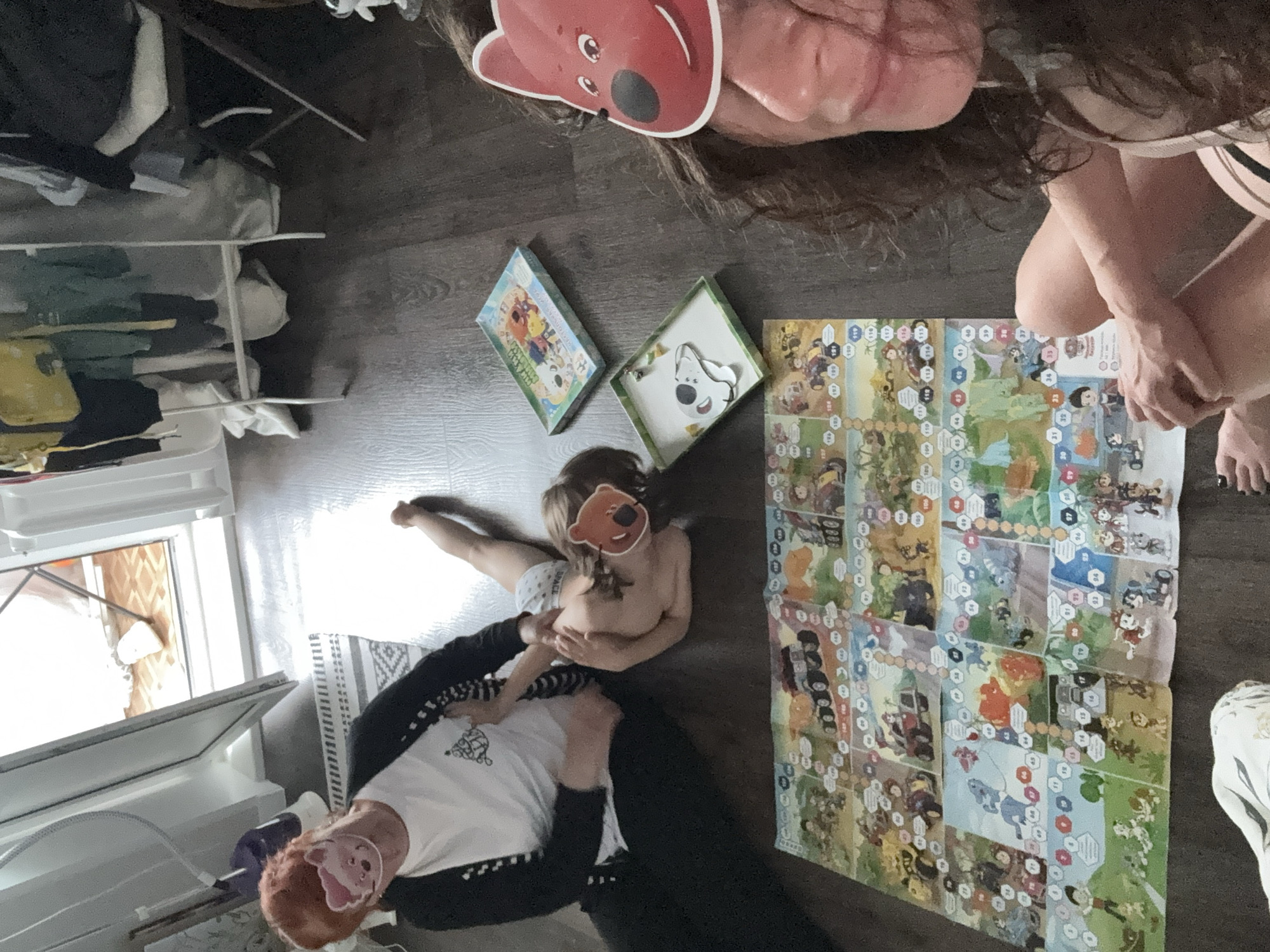 In an indoor setting, likely a home in Chaikovsky, Russia, two individuals are playing a board game on a dark wooden floor. One person, an adult, is wearing a white t-shirt with a black cardigan and black pants. The other person, a child, is lying on their side, wearing only underwear. Both are wearing animal-themed masks over their faces. The board game is laid out on the floor and features colorful illustrations and hexagonal spaces. Two game boxes are also visible nearby. In the background, a white clothing rack is filled with clothes, and a window with an open sill shows a glimpse of the outside. The overall scene suggests a casual and playful family activity.