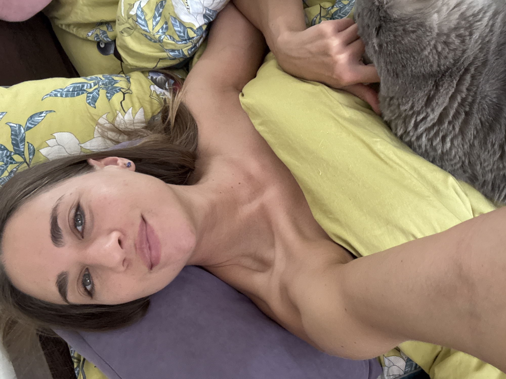A young woman with brown hair is lying in bed. She is looking at the camera with a slight smile. The woman is wearing a light-colored top that is off her shoulders. She has a purple pillow behind her head and a yellow pillow under her arm. A gray cat is lying next to her. The room is decorated with floral patterns on the bedding. The scene is a bedroom in a home in Unknown, Russia.