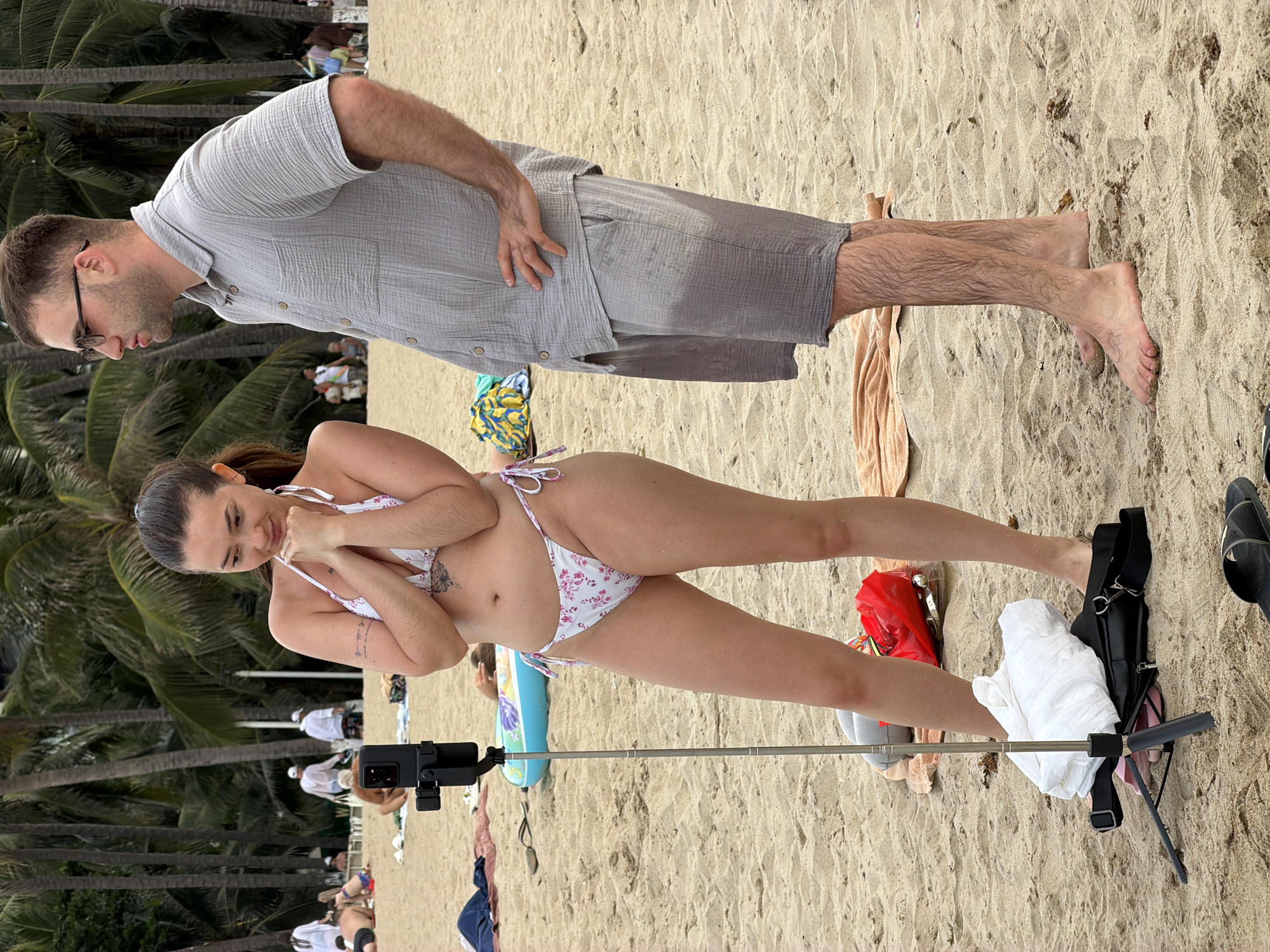 In Nha Trang Ward, Vietnam, on a sandy beach, a young woman in a floral bikini poses with her hands clasped near her chin, looking down and to her left. Beside her stands a man wearing a gray, short-sleeved button-up shirt and gray shorts, with his right hand on his hip and looking to his left. A black selfie stick with a phone attached is positioned in front of the woman on the sand. In the background, palm trees line the beach, and other beachgoers are visible, some reclining on towels or walking. The environment is a typical beach scene with sand, water in the distance, and a generally overcast sky.