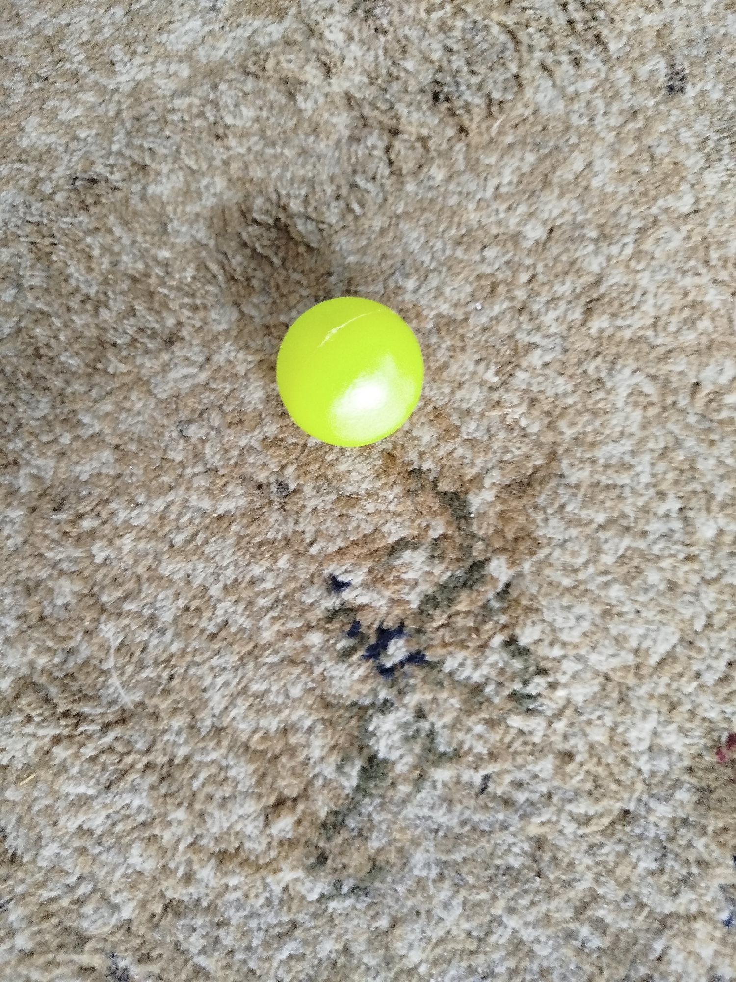 A single, bright yellow, smooth ball rests on a textured, light brown carpet. The carpet appears to be woven with varying shades of brown and white fibers, creating a fuzzy, almost shag-like appearance. There are a few darker specks and patches visible within the carpet, suggesting dirt or shadows. The lighting in the image is direct, causing a slight shine on the surface of the ball and casting subtle shadows on the carpet beneath it. The ball is positioned slightly off-center towards the top of the frame. The scene is devoid of any human presence or discernible urban environment. The specific location in Крывасельскі сельскі Савет, Belarus cannot be determined from the provided image.