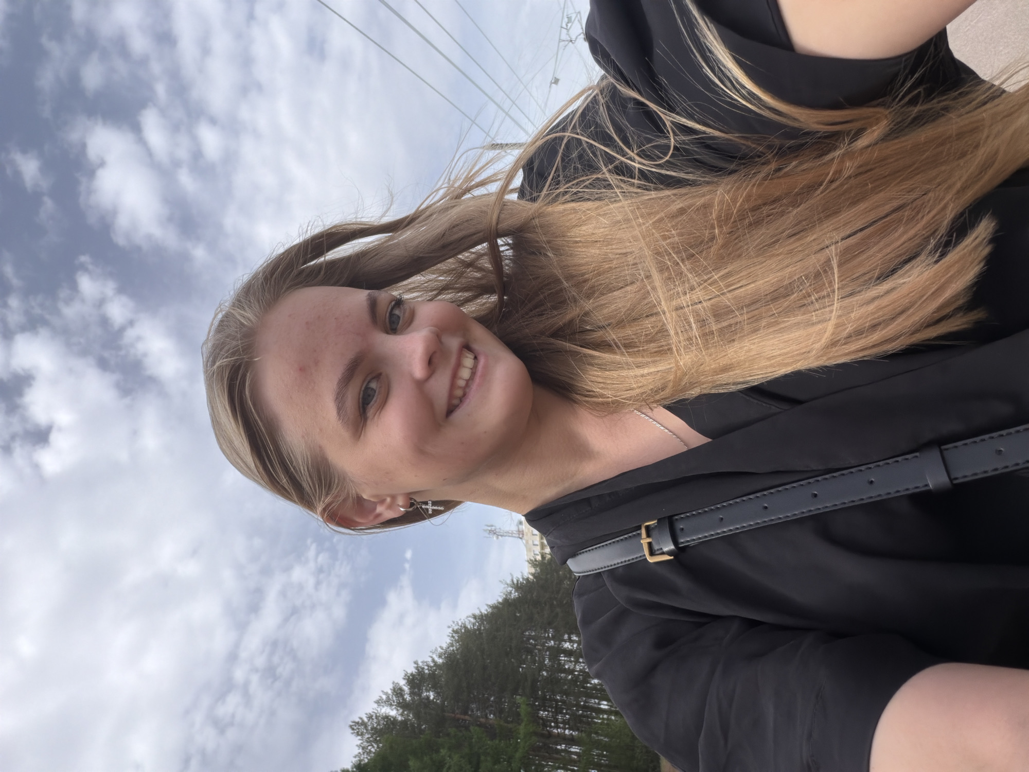 A blonde-haired female is in the city of Izhevsk, Russia. She is seen from the chest up in a selfie style and is smiling with sunlight on her face. She has a black crossbody purse and a black top. There is a cloud-filled sky, power lines, a radio tower, and trees behind her.
