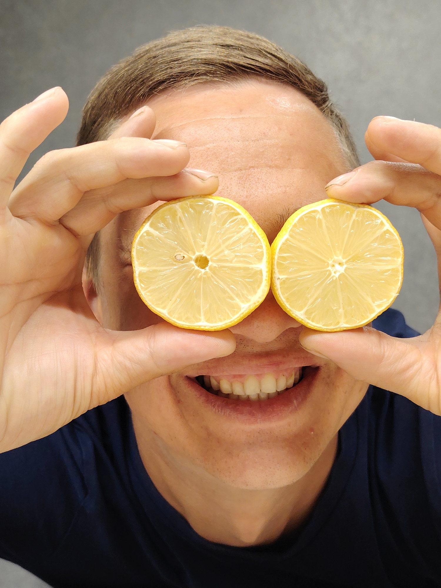A close-up, head-and-shoulders shot shows a person in Chaikovsky, Russia, holding two lemon slices over their eyes, obscuring them. The person is smiling broadly, with their mouth open, revealing teeth. Their hands are visible on either side of their face, framing the lemons. They are wearing a dark blue t-shirt. The background is a plain gray wall. The lighting appears to be artificial, with no indication of time of day or weather. The image is candid, conveying a playful or humorous emotion. No text is visible in the image.