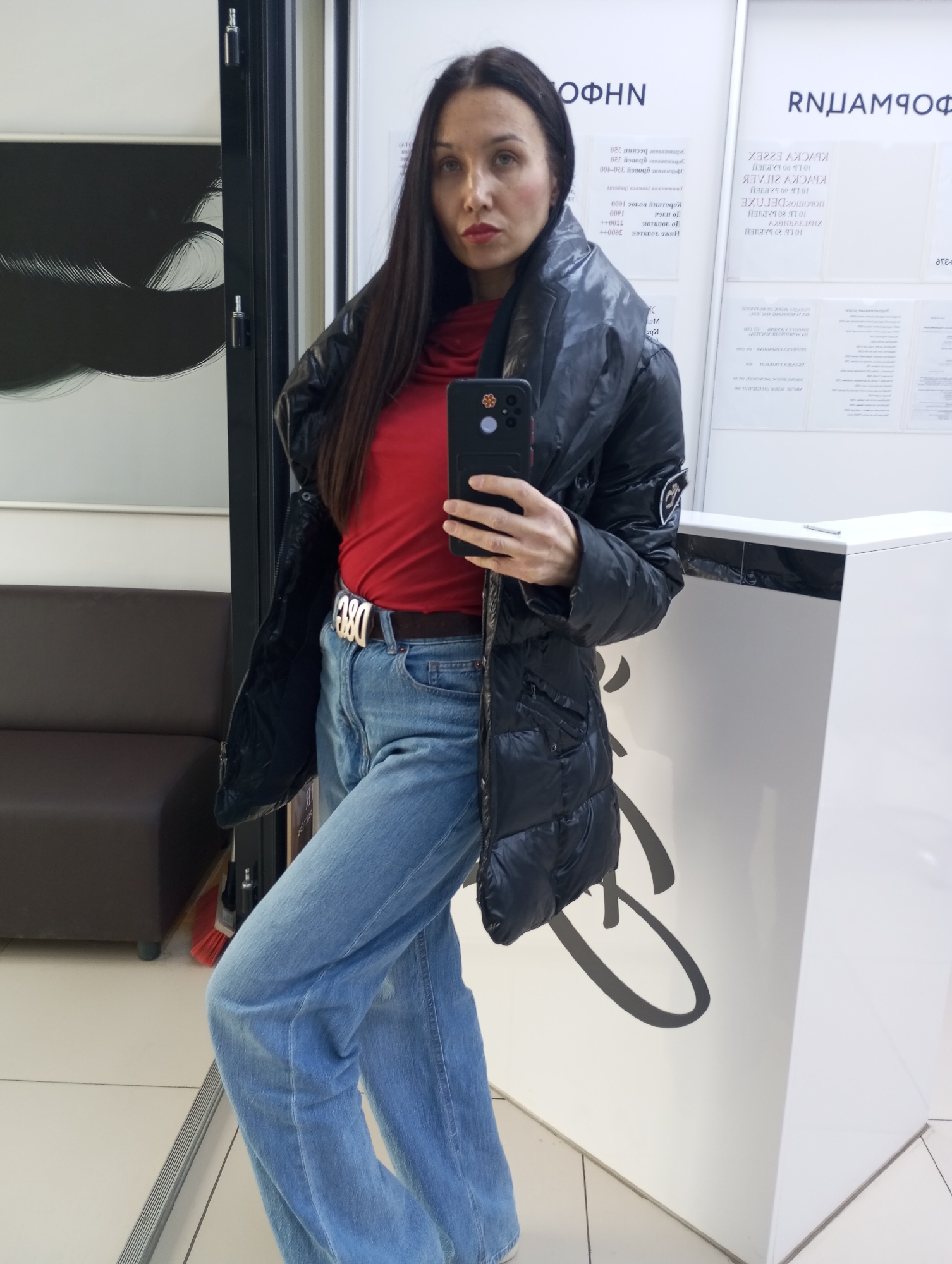 A person is captured in a full-length mirror selfie within an indoor setting, likely a commercial or public space in Izhevsk, Russia. The subject is a woman with long, dark hair, wearing a black puffer jacket over a red turtleneck or high-necked top, and light blue wide-leg jeans. A prominent belt with a metallic "D&G" buckle is visible around her waist. She holds a black smartphone with a case in her right hand, positioned to take the photo, and looks directly towards the camera with a neutral expression.
The background shows reflections typical of a mirrored environment. To the left, a dark brown upholstered couch is visible, and below it, a red broom. The wall behind the subject and to her left features a black and white abstract design. To her right, a white counter or reception desk is visible. The white walls in the background reflect various pieces of text, indicating information or advertisements, some of which are clearly mirrored.
Visible mirrored text includes "RNUAMIOФ", "ΝΗΦΟ", "КБУСКУ ГІГЛЕК", "КЬУСКУ Е22EX", "ПОБОТОК ДЕГОХЕ", "10 15 00 ΚΛΕΙEN", "ΑΧΘΗΒΑΣ ΜHX", "10 15 20 БАРИEN", along with numbers and other fragmented words like "320", "1000", "3300++", and "CATO", which appear to be related to prices or services. The overall impression is a casual pose in a functional, well-lit interior. The time of day is not discernible.