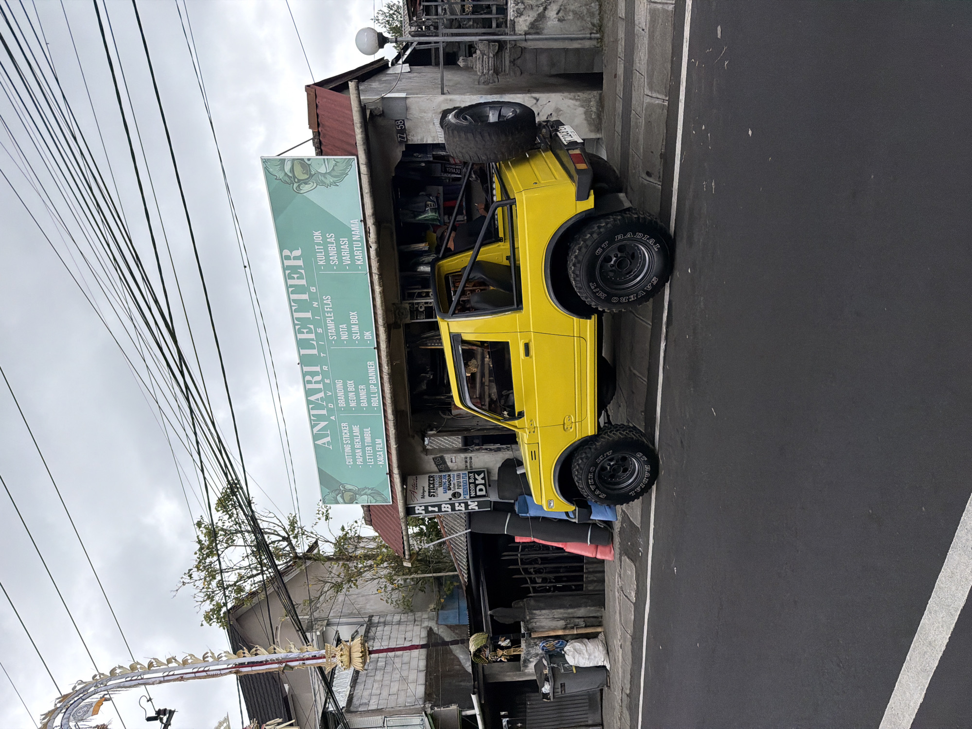 An overcast day in Kintamani, Indonesia, shows a bright yellow, open-top Suzuki Samurai jeep parked on the side of a paved road. The jeep is facing left, with its rear tire prominently displayed on a spare mount at the back. In the background, a building with a green sign that reads "ANTARI LETTER ADVERTISING" is visible. The sign features a cartoon drawing of a monkey wearing sunglasses. The building appears to be a storefront, with products or materials displayed behind a glass window. To the left of the storefront, a tall, ornate Balinese ceremonial banner (umbul-umbul) stands. The street in front of the building has a white line marking the edge of the road. Several large rolls of material, possibly for signage or printing, are stacked vertically next to the storefront. Trash bins and bags of waste are also present on the sidewalk. The environment suggests an urban or semi-urban setting, possibly a commercial area.