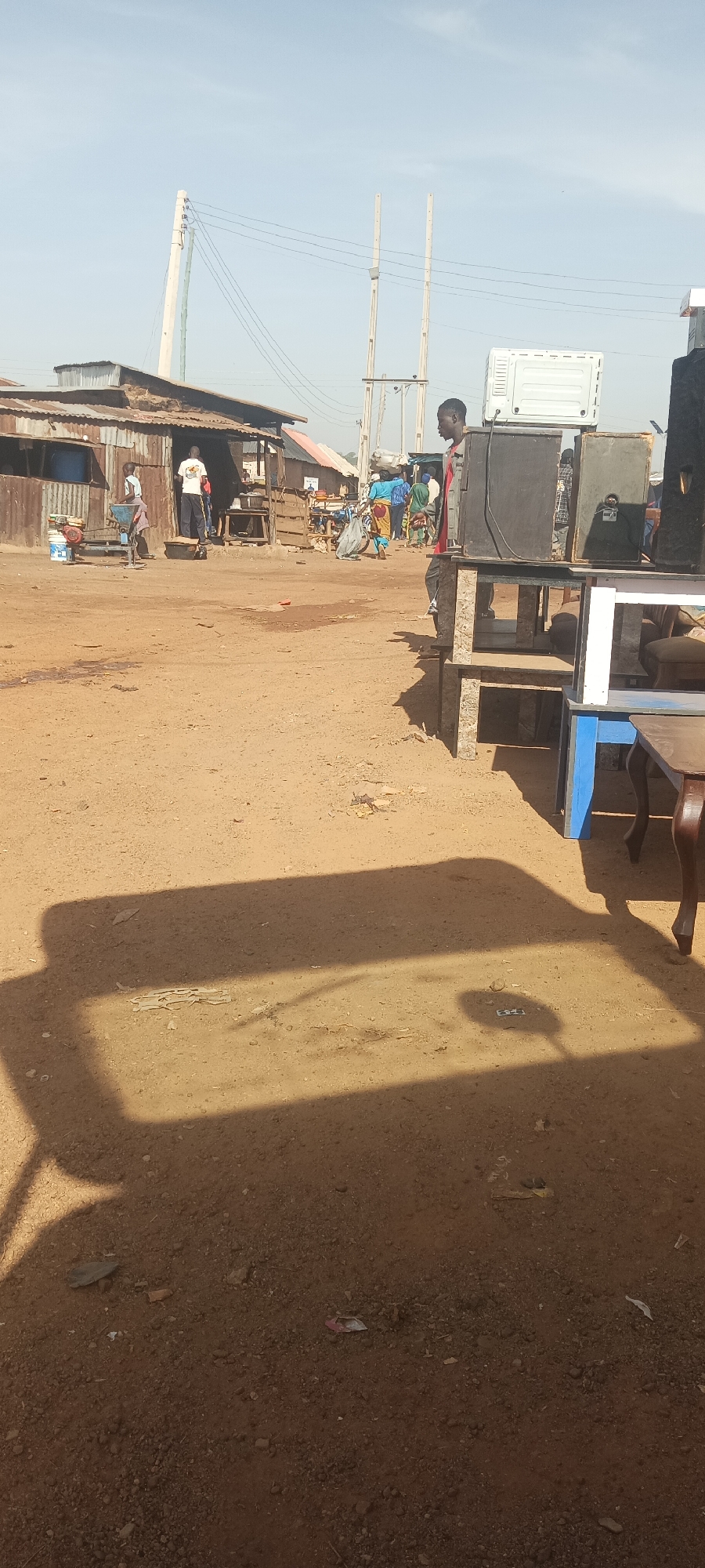 This outdoor image captures a bustling scene, likely a market or street in a semi-urban or rural area, under a bright, hazy blue sky. The ground is unpaved, consisting of reddish-brown dirt and sand, with scattered debris. Long, distinct shadows are cast from the right side of the frame towards the left, suggesting either early morning or late afternoon sun.
In the immediate foreground on the right, several large, dark-colored speakers or sound system components are stacked on simple stands made of concrete blocks. Adjacent to these are various wooden tables, including one with bright blue painted legs and another with intricately carved, brown wooden legs, implying they are items for sale or display. A man in a red shirt stands facing left in the mid-ground, positioned beside these items. Behind him, a white rectangular object, possibly a large appliance like an air conditioner unit, is visible.
In the left background, several informal structures with corrugated metal roofs and walls line what appears to be another street or pathway. People are visible near these buildings; one person in a white t-shirt stands near a structure, while another operates a blue and white machine on the ground. Further in the background, more figures can be seen, with at least one person carrying something balanced on their head.
Above the buildings and people, tall utility poles, appearing to be concrete pillars, support numerous power lines that stretch across the sky. The overall impression is one of daily life and commerce in a lively, informal setting. No discernible text is clearly visible on any objects or clothing.