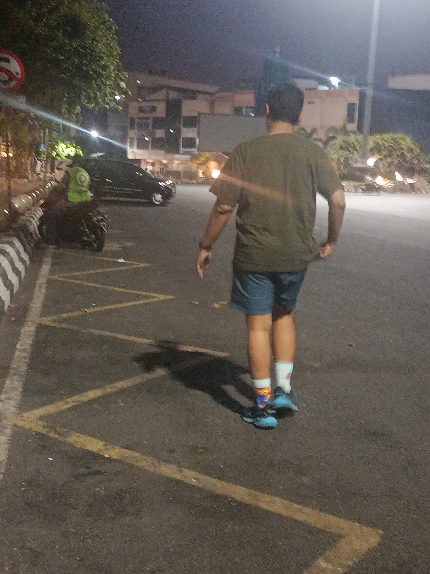 A dimly lit, nighttime street scene in Semarang, Indonesia, captures a person walking away from the camera. The individual, wearing a khaki-colored t-shirt and blue shorts, has brightly colored socks and blue sneakers. A black motorcycle is parked on the left side of the frame, with a person in a bright green vest standing near it. A black car is also visible in the background, parked behind the motorcycle. The street is marked with yellow lines, indicating parking spaces or traffic lanes. Trees and a building with illuminated windows are present in the background, suggesting an urban environment. The overall atmosphere is dark and slightly hazy, with some light sources creating glare and reflections on the wet asphalt.