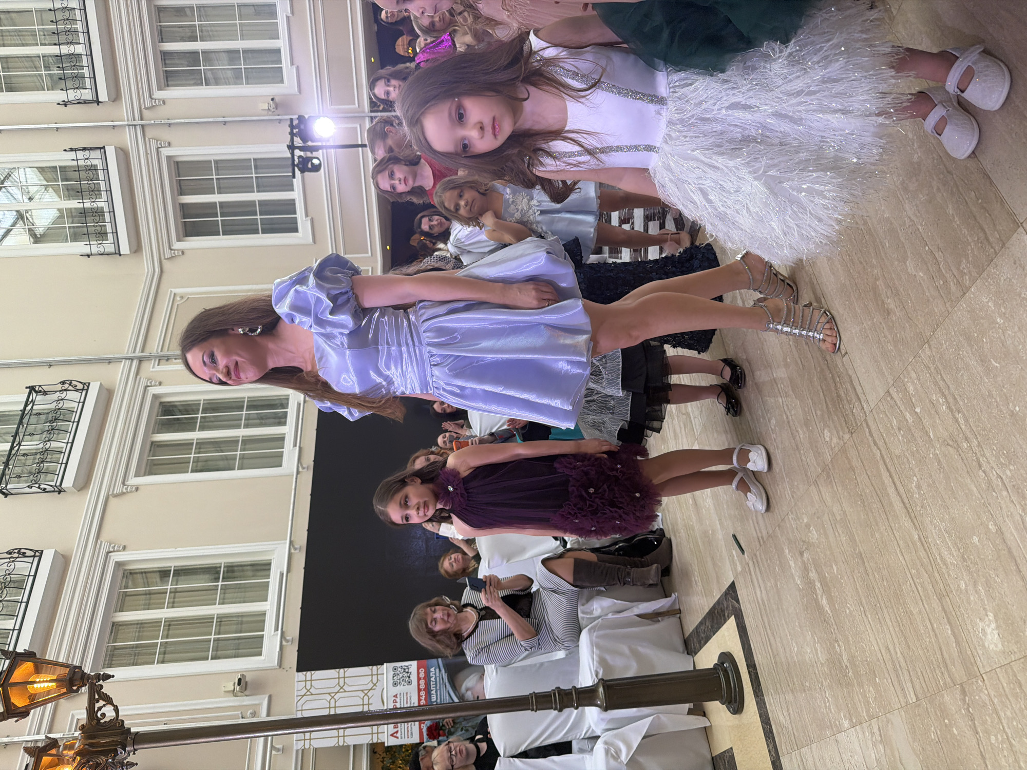 The image shows a fashion show, possibly for children's clothing, taking place in what appears to be an indoor space in St. Petersburg, Russia. Several girls and women are posing, wearing formal dresses. One woman in a light purple dress and silver strappy heels stands prominently. The backdrop features a building facade with windows. Attendees are seated in the foreground. A lamp post with an ornate, multi-bulb fixture is visible on the left. The QR code is readable. The floor is tiled.