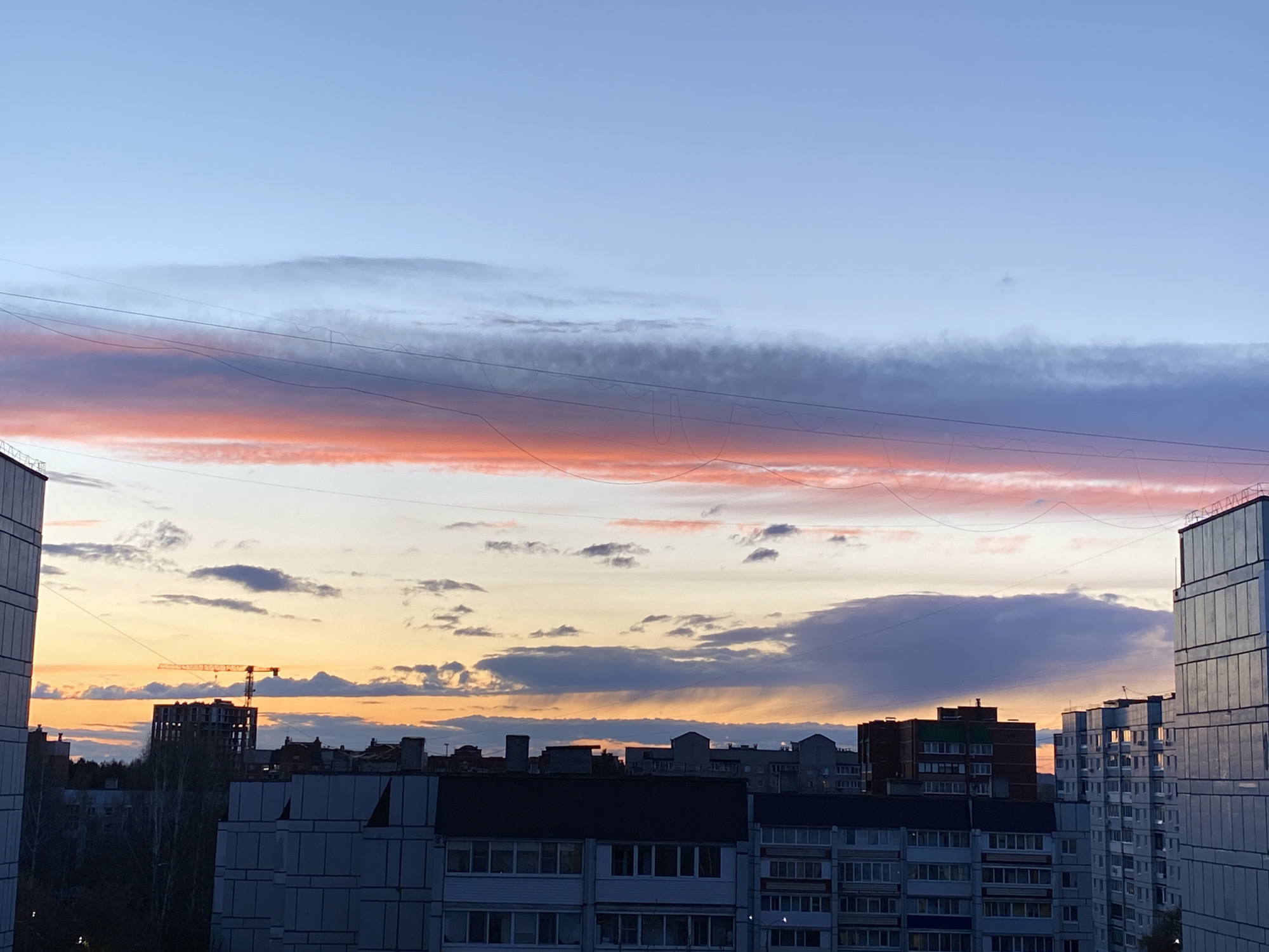 The image captures an urban skyline in Chaikovsky, Russia, at either sunrise or sunset, characterized by a dramatic and colorful sky. The upper sky is a clear light blue, transitioning downwards into a prominent horizontal band of vibrant pink and orange clouds, suggesting the intense hues of the rising or setting sun. Below this, cooler grey and blue clouds are visible, interspersed with patches of lighter orange and yellow closer to the horizon line.
Several dark, thin utility wires crisscross horizontally across the mid-upper portion of the sky, adding a linear element to the natural backdrop.
The foreground and mid-ground are dominated by various multi-story urban buildings. On the far left, part of a tall, grey building is visible. In the center, multiple apartment-style buildings with numerous windows, some light-colored and some with darker upper sections, form a dense urban fabric. To the far right, a modern-looking, tall building with a reflective glass facade stands out. In the mid-distance, a dark-colored building under construction, made primarily of concrete, is topped by a large yellow tower crane, signifying ongoing development. Dark, bare tree branches are visible in the gaps between some buildings.
No people or explicit activities are depicted, making it a serene landscape view of the city. The low angle of the sun casts the buildings in soft light or silhouette, enhancing the vividness of the sky. No visible text is present in the image.
