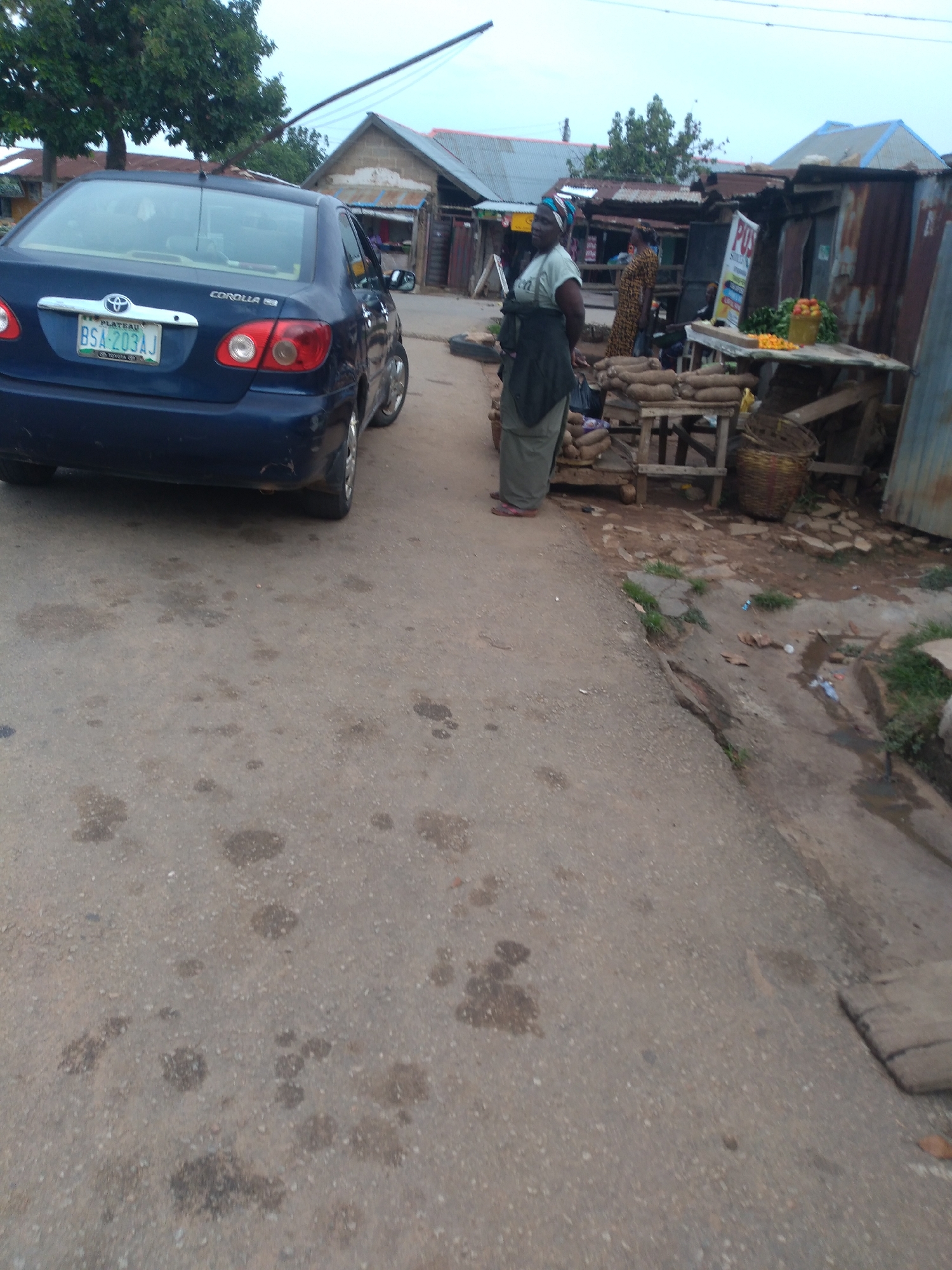 The image depicts an outdoor scene, likely a roadside market or commercial area, in Jos, Nigeria. A dark blue Toyota Corolla is parked on the left side of a paved road, its license plate reading "BSA-203AJ". The road surface is weathered and shows dark patches, possibly oil or water stains.
On the right side of the image, a woman wearing a headscarf and a green dress stands near a market stall. She is facing away from the camera, observing the scene. Behind her, another woman in a patterned dress is visible, also near a stall.
The market area features various stalls constructed from corrugated metal and wood. Some stalls display produce, including what appears to be yams piled on wooden stands. A woven basket sits on a wooden structure. A sign with the acronym "POS" is prominently displayed on one of the stalls. The environment appears somewhat unkempt, with debris and vegetation visible on the ground, particularly in the drainage area on the far right. The sky above is overcast.
