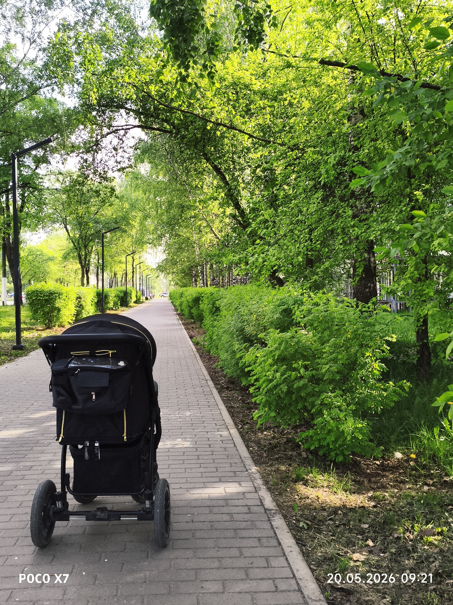 A stroller stands on a brick-paved path in a park or green area, likely in Chaikovsky, Russia, as indicated by the surroundings. The path stretches into the distance, lined with green bushes and trees, forming a canopy above. Lampposts are visible along the path. The stroller is black, with a visible bag or storage compartment attached. The date and time, "20.05.2026 09:21," are stamped on the lower right of the image, and "POCO X7" is on the lower left, suggesting the image was taken with a POCO X7 phone. The overall scene suggests a calm, potentially early morning stroll. There are no people visible in the frame.
