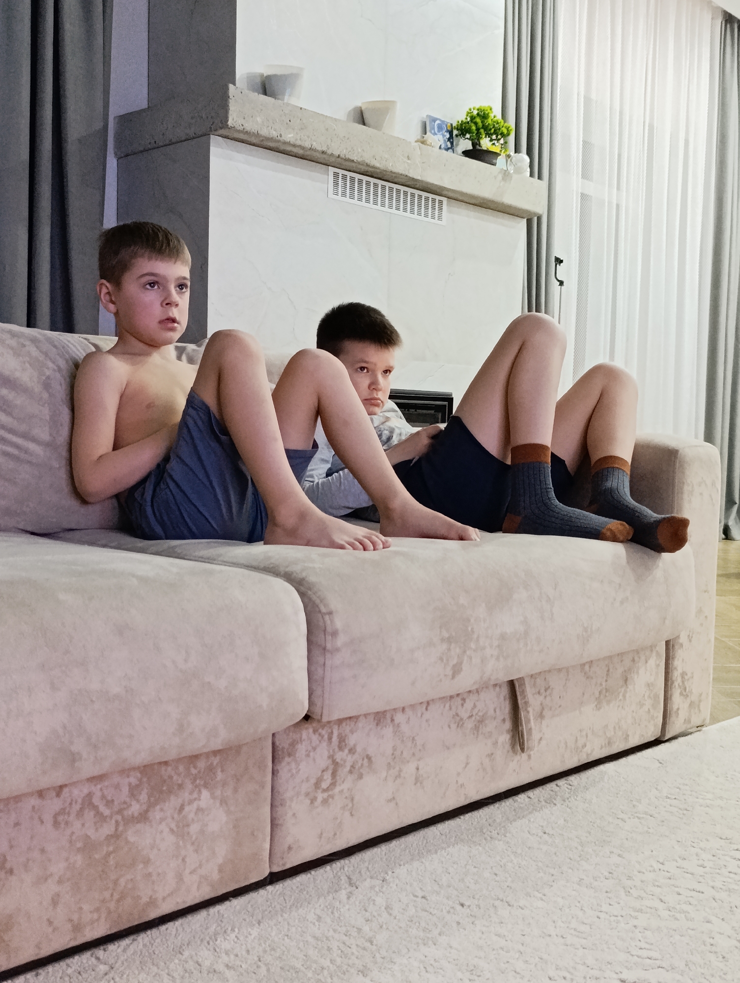 Two boys sit on a light-colored sofa. The boy on the left is fair-haired and shirtless, wearing blue shorts. He stares ahead. The boy on the right has dark hair and wears a light-colored shirt with blue shorts. He has dark gray socks with brown heels and toes on his feet. The boys are situated in front of a fireplace with a white mantelpiece and a decorative shelf holding plants and vases. Gray curtains are partially visible in the background. A light-colored carpet covers the floor. The location is the city of Chaikovsky, Russia.