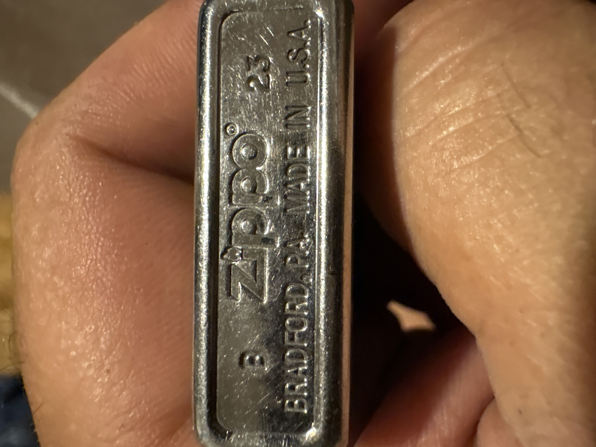 The image shows a close-up of a Zippo lighter held by a hand. The lighter is engraved with the Zippo logo, the text "BRADFORD, PA. MADE IN U.S.A.", and what appears to be a date code "23" and a letter "B". The background is blurred and consists of skin texture and possibly fabric. The image does not contain any information about the city of Hradec-Nová Ves, Czechia.