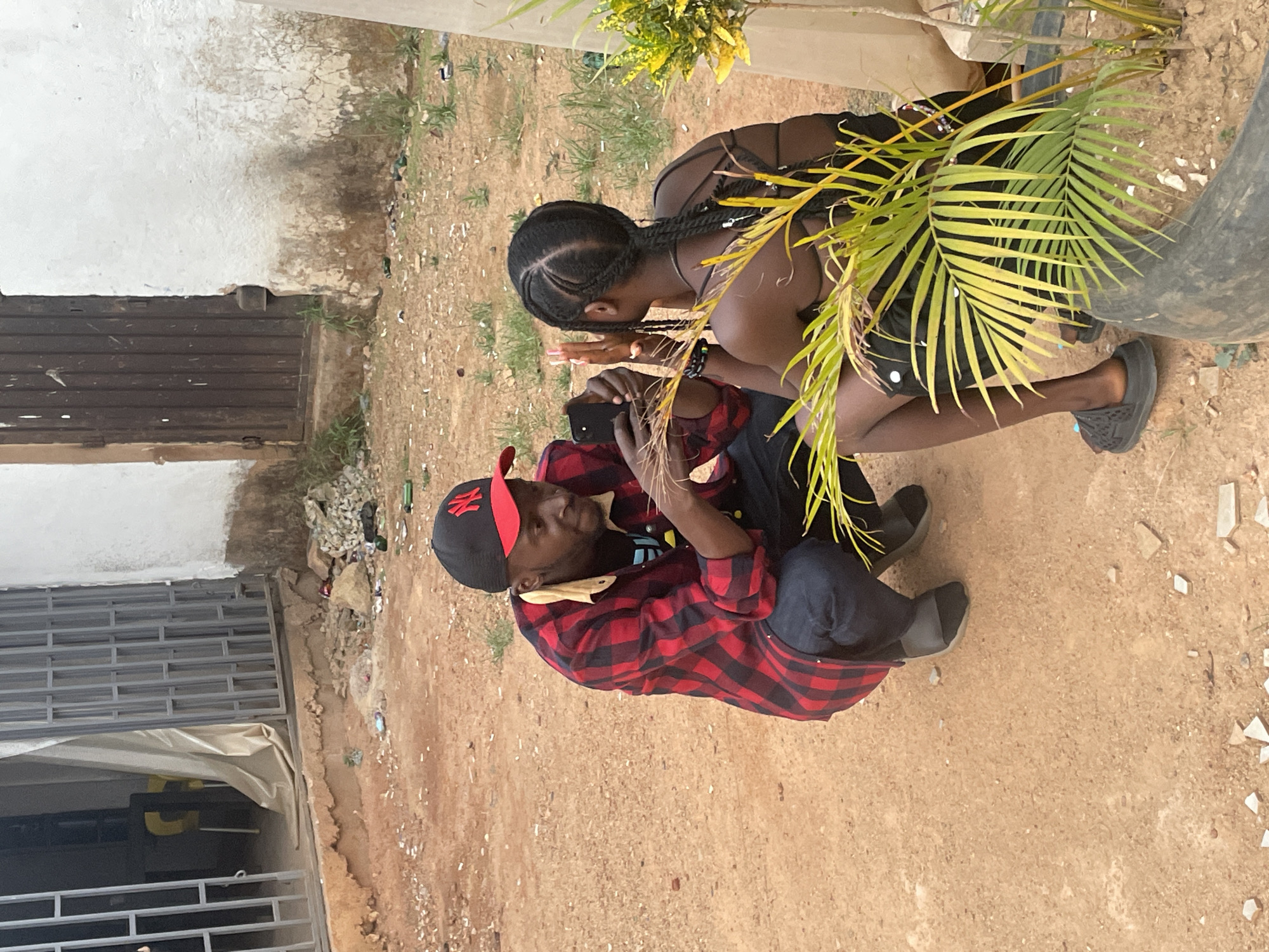 Two individuals are positioned outside, potentially in Barakin Akawo, Nigeria. A man wearing a red and black checkered shirt, a black baseball cap with a red logo, and dark jeans is squatting while holding a black smartphone in his hands. He appears to be looking at the phone. Next to him, a woman wearing a black top and shorts is also squatting, seemingly interacting with the man and his phone. A green, leafy plant obscures part of their interaction. The ground appears to be dirt or sand. The edge of a building with a barred window is visible to the left. There is some trash scattered on the ground.