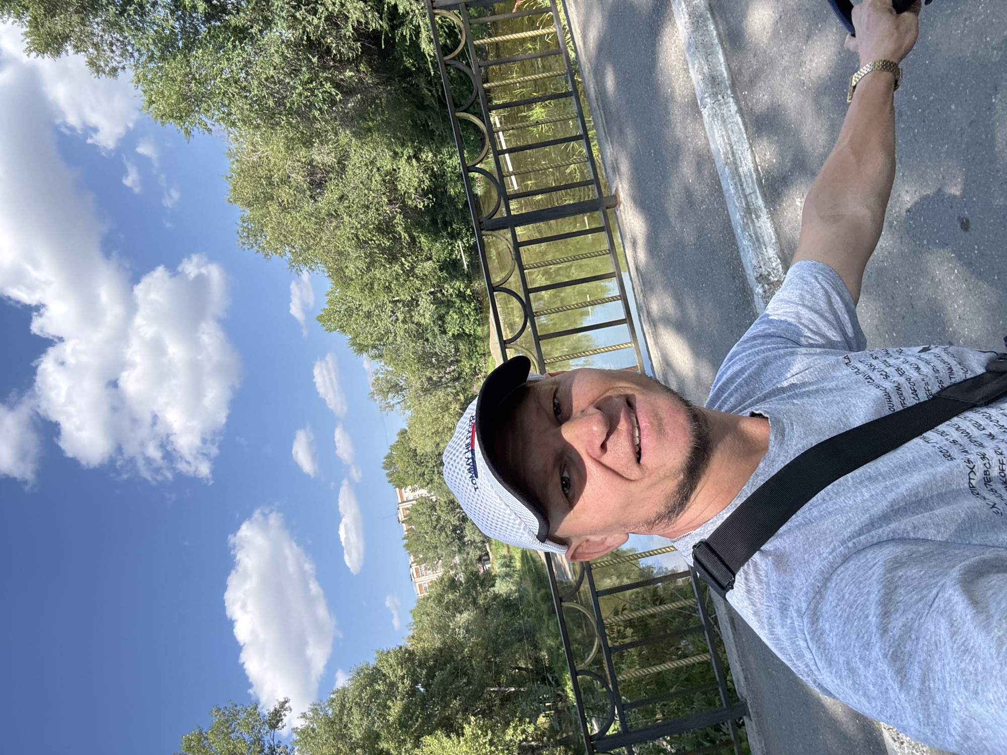 A man wearing a grey t-shirt, a black strap across his chest, and a white Tommy Hilfiger baseball cap is taking a selfie on a bridge. The bridge has a black metal railing with decorative gold accents. Beyond the railing is a body of water with vegetation growing in it, reflecting the sky and trees. The background shows a dense line of green trees, with a multi-story building visible through a gap in the trees. The sky is blue with scattered white clouds. The man's right arm is visible, resting on what appears to be a bicycle handlebar, and he is wearing a gold watch. The image appears to be taken during the daytime with bright sunlight casting shadows. The bridge and surrounding scenery are consistent with the location of the Imannov bridge in Oskemen, Kazakhstan, which spans the Irtysh river and is a popular spot for pedestrians and cyclists.