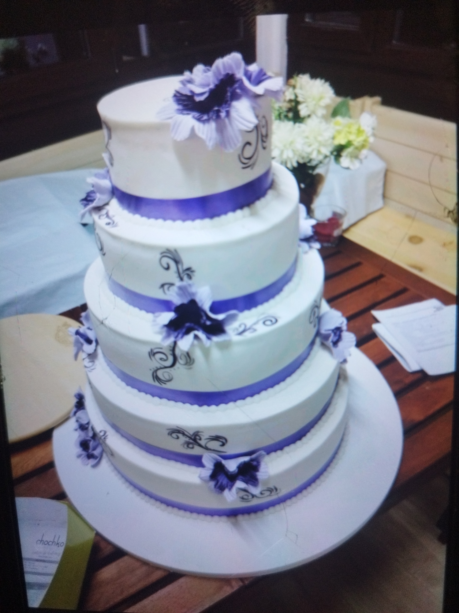 A four-tiered white cake decorated with purple ribbon, purple flowers, and black scroll designs is the focal point of the image. The cake sits on a white circular base. A card with the name "chochko" visible is on the lower left side of the cake. A small arrangement of white and yellow flowers in a clear vase is behind the cake. The scene is set on a wooden table with a slatted design. Loose papers are on the table's right side. The location and city are not evident in the image.