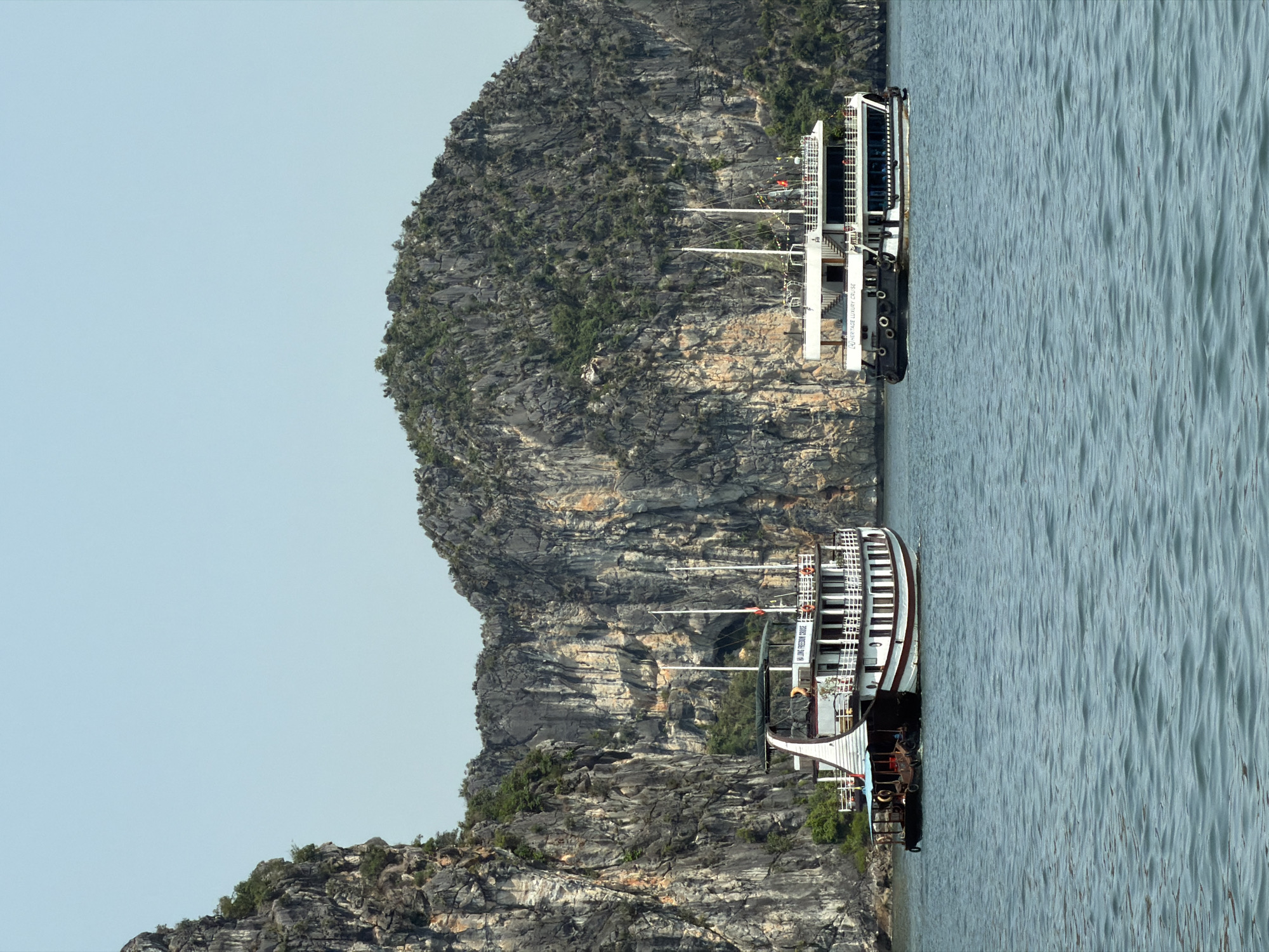 The image displays a picturesque scene of two cruise boats navigating a calm bay, flanked by majestic limestone karsts, under a clear, bright sky. The photograph is taken during the daytime, with ample natural light illuminating the landscape.
In the foreground, a multi-decked cruise ship, predominantly white with dark hull accents, glides across the water. It features several levels with visible railings and windows, and text on its side clearly reads "HALONG FREEDOM CRUISE". Two tall, slender masts or poles rise from its deck.
Further in the midground, another similar cruise vessel is visible, partially silhouetted against the karsts. This boat is also white, multi-tiered, and has "HERITAGE LUXURY CRUISE" inscribed on its side. Both boats appear to be luxury or tour operators.
The background is dominated by towering, rugged limestone karsts, typical of a dramatic coastal landscape. These islands are covered with patches of lush green vegetation clinging to their steep, grey and tan rock faces. The water in the bay is a tranquil greenish-blue, showing subtle ripples on its surface, indicating calm conditions. The sky is a uniform, pale blue, suggesting fair and clear weather. The scene captures a serene moment of cruising amidst the natural beauty of Tuan Chau Ward, Vietnam.