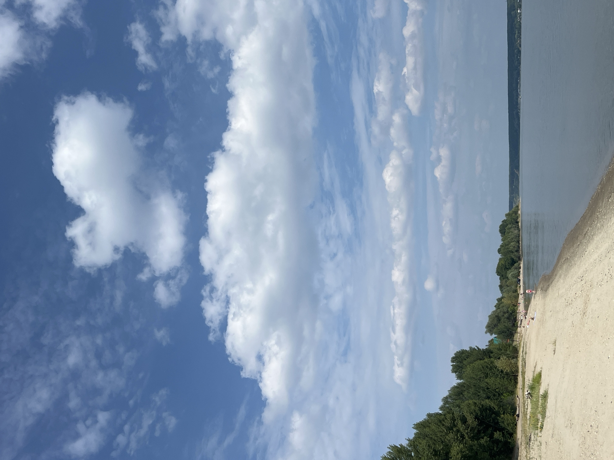 The image depicts a wide, calm river with a sandy beach along its left bank. The beach is lined with a dense forest of green trees, with a few scattered structures visible amongst the foliage. A number of people are present on the beach, some walking and others relaxing. The sky is a bright blue, filled with scattered white clouds, suggesting fair weather. The scene is likely a popular spot for recreation and relaxation during the summer season. The location is identified as Mochishche, Russia.