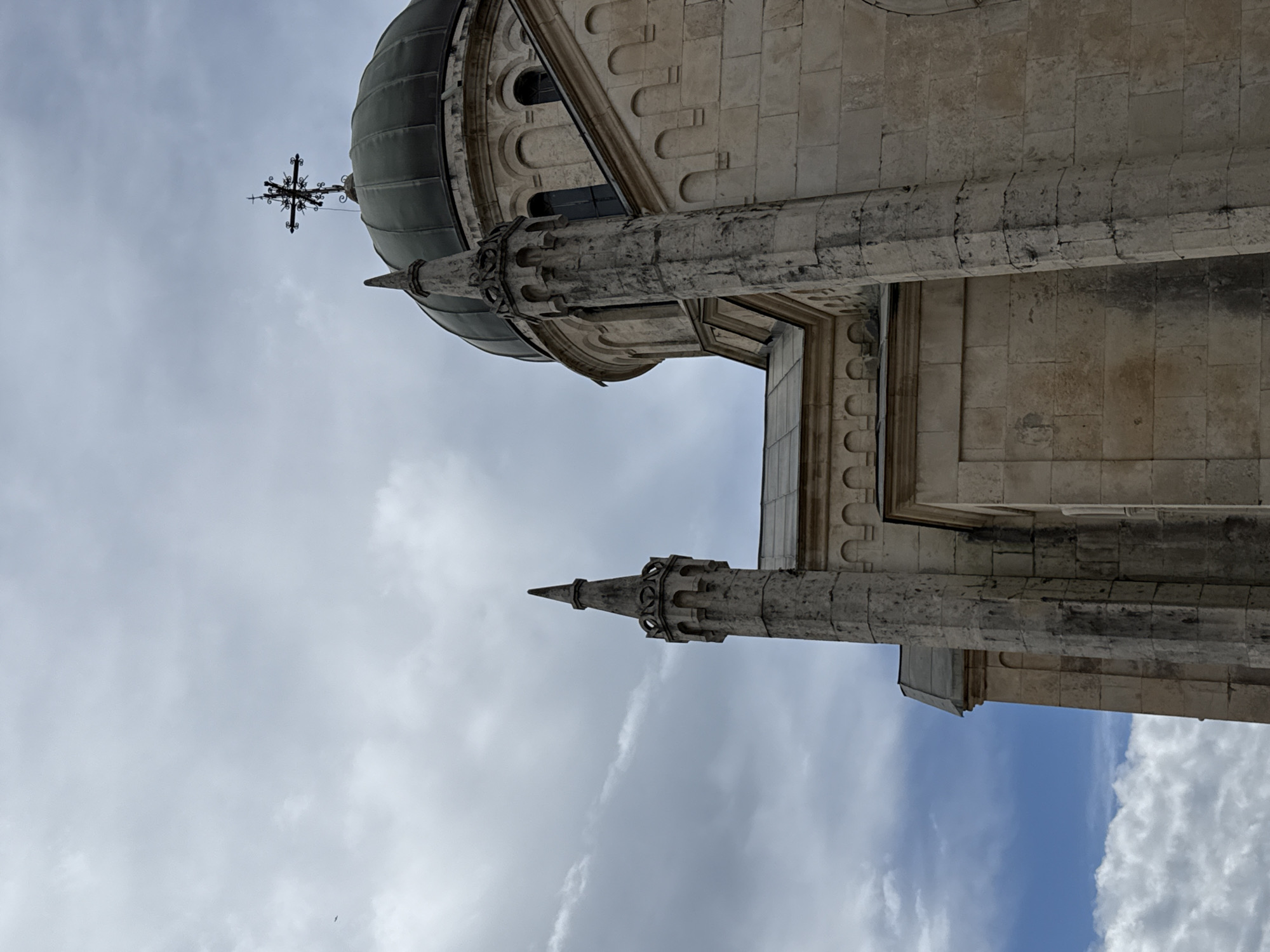 The image shows the Savina Monastery in Herceg Novi, Montenegro. The scene captures an upward angle of the building's exterior, focusing on the dome with a cross on top, a tower-like structure, and parts of the stone facade. The sky with clouds is visible in the background.