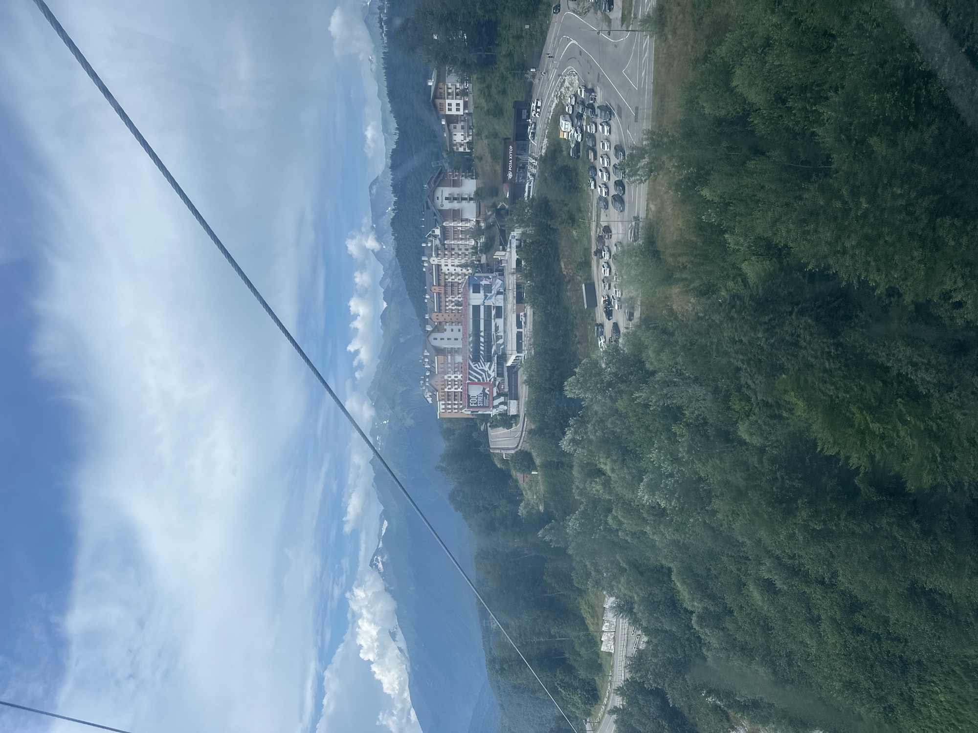 This is a telephoto image taken from a cable car looking down on the ski resort area of Rosa Khutor in Estosadok, Russia. The scene depicts a mountain landscape with a village nestled in the valley below. In the foreground, a dense forest of green trees covers the slopes. Above the trees, a multi-lane road curves through the landscape, with numerous cars parked on either side of it, indicating a busy area. Several modern buildings, characteristic of a ski resort, are situated on a hillside. These buildings appear to be hotels or apartment complexes, some with balconies and a mix of white and brown facades. One prominent building has a large graphic advertisement on its side. Further in the background, majestic, snow-capped mountains rise against a partially cloudy sky. The cable car's wire is visible in the upper portion of the image, stretching diagonally across the frame, suggesting the perspective of the viewer. The overall impression is one of a well-developed tourist destination situated in a dramatic natural environment.