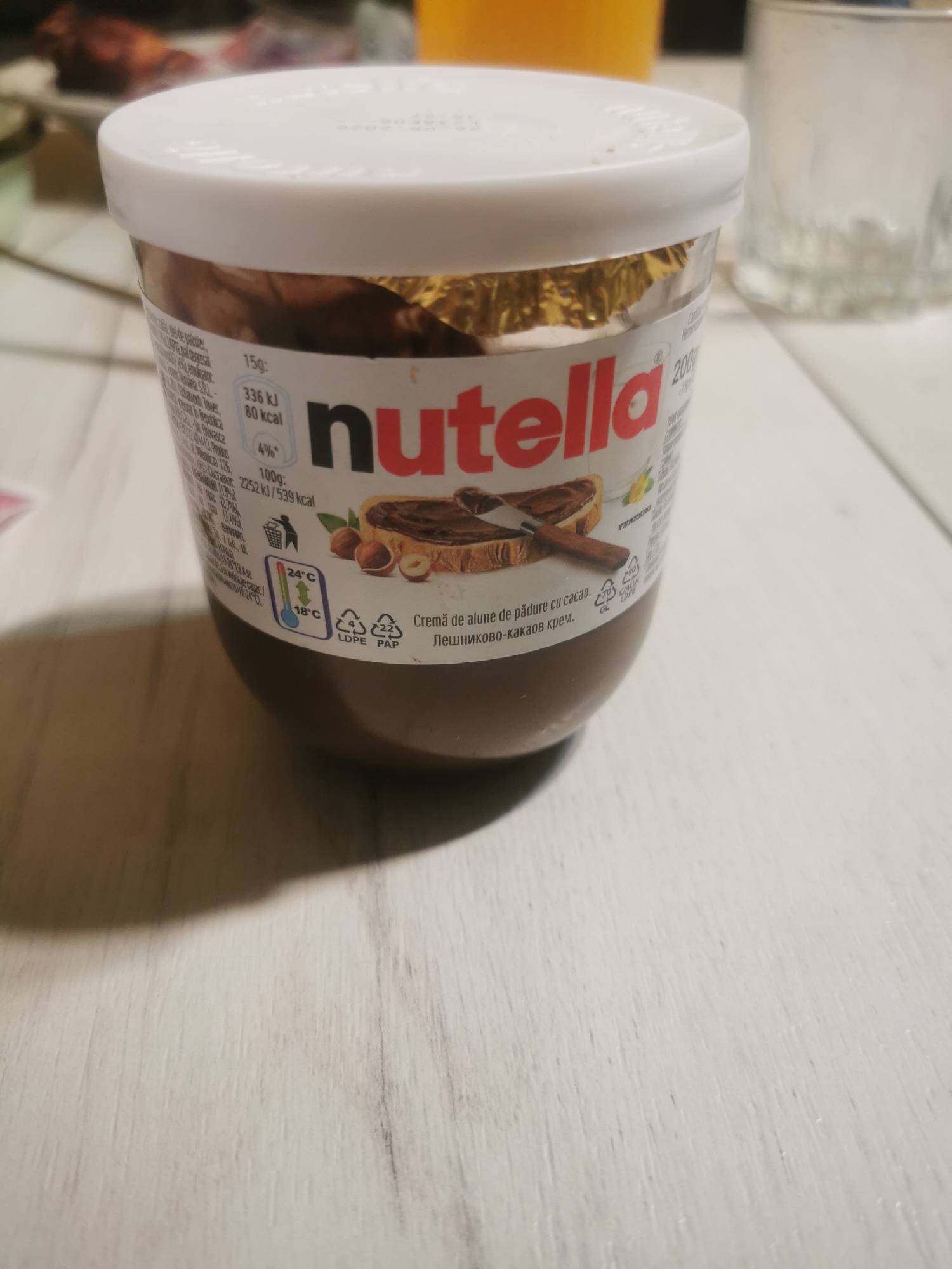 The image shows a jar of Nutella hazelnut spread with cocoa. The label is partially visible, displaying nutritional information in multiple languages, including Romanian and Bulgarian. The jar is set on a white surface, possibly a table or counter. The environment appears to be indoors.