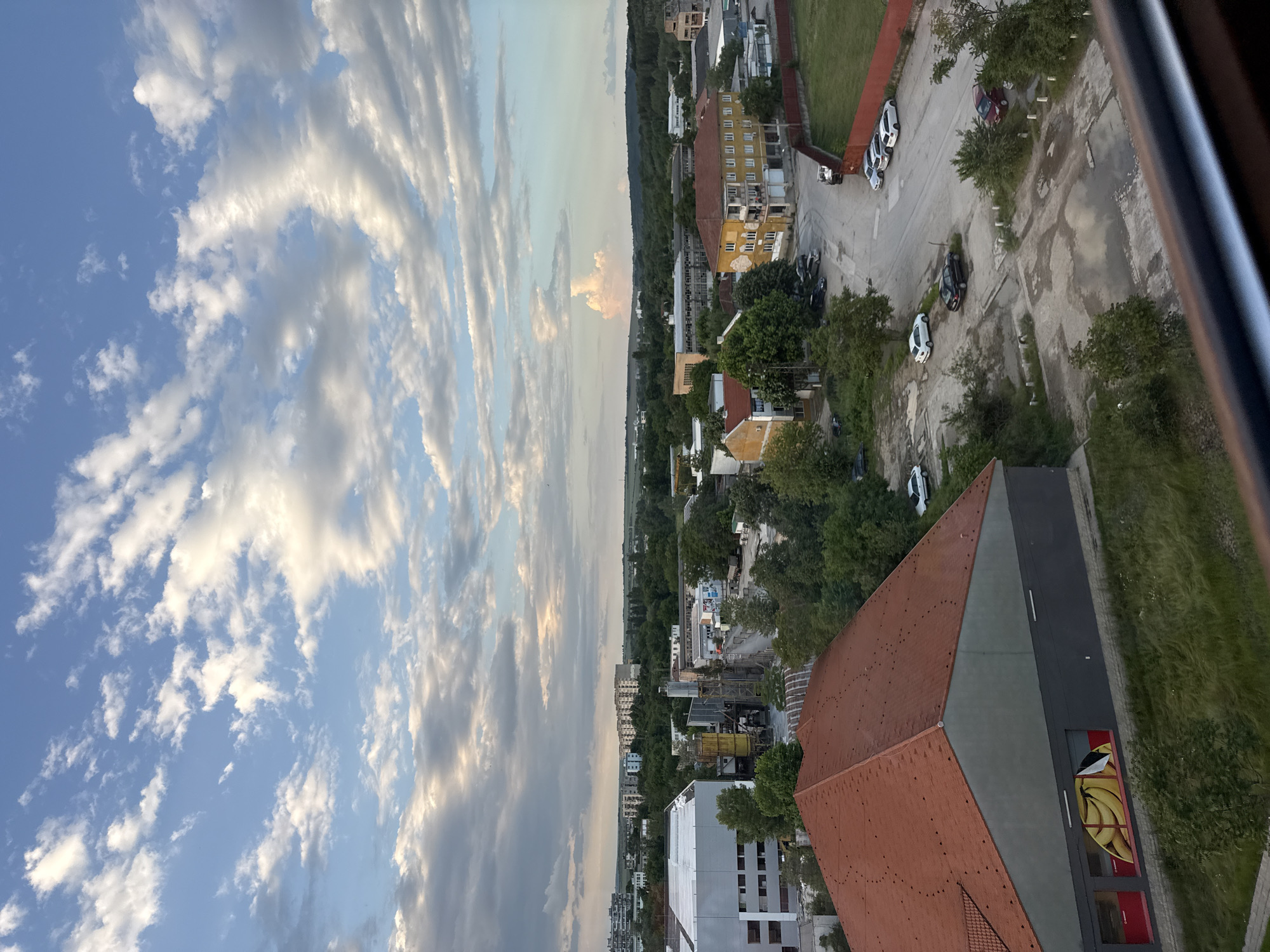 The image is an aerial view of a cityscape in Lovech, Bulgaria, captured from a high vantage point, possibly a building or elevated structure. The sky dominates the left side of the frame, showcasing a mix of blue sky and wispy, elongated clouds. The city itself is visible on the right, consisting of buildings of varying sizes and styles, interspersed with green spaces and trees. Some buildings have orange roofs, while others are white or gray. A street runs through the urban area, with several parked cars visible along its side. A distinctive building with a red-tiled roof and a grey wall with a mural of bananas is prominent in the foreground. The overall impression is of a clear day with a mix of residential and commercial areas in the city.
