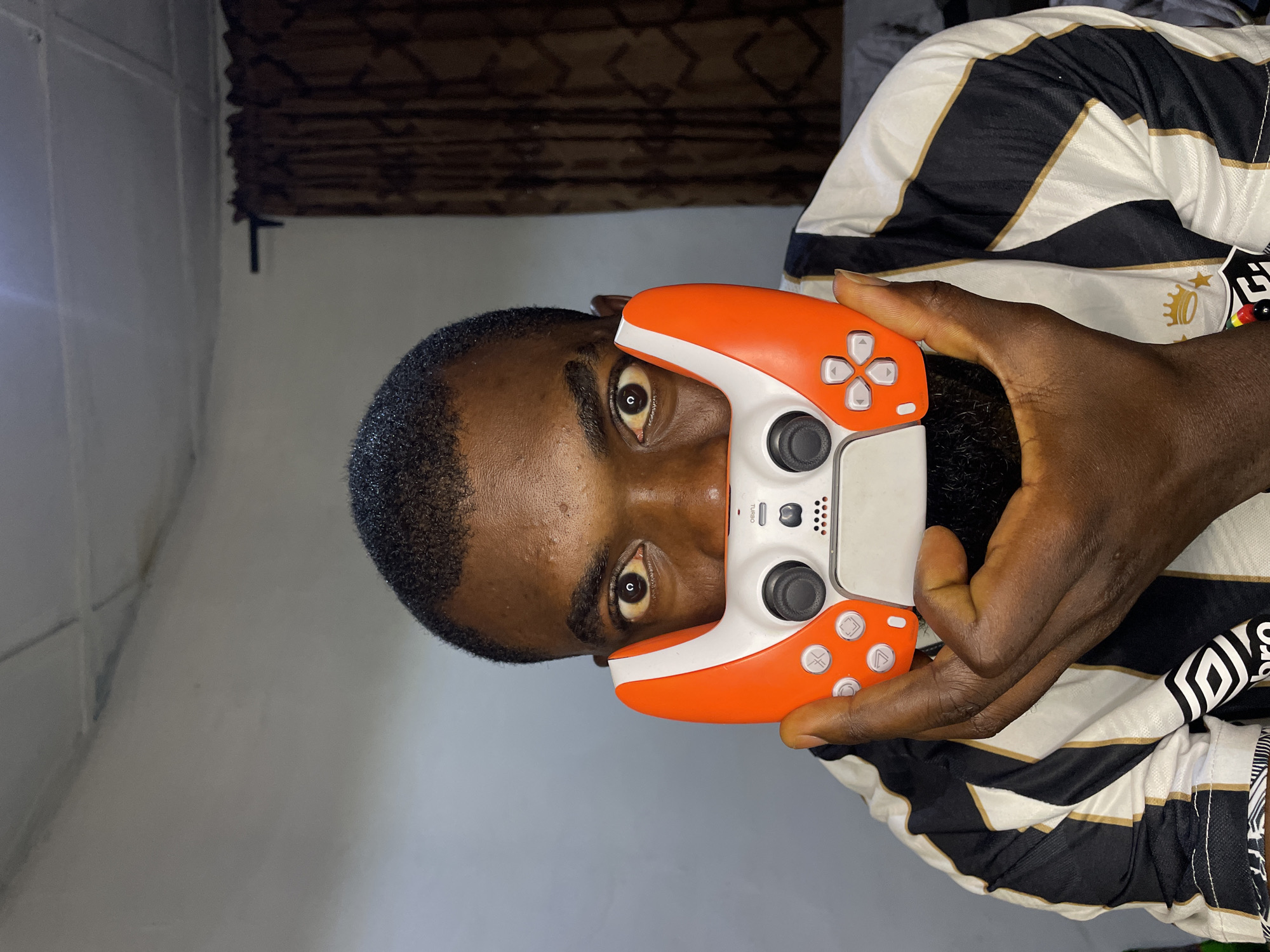 A close-up portrait shows a Black man's face and hands in the foreground. The man, who is wearing a black and white striped shirt, is holding an orange and white video game controller to cover the lower half of his face. His eyes are wide and looking directly at the camera. The background is dimly lit, with a textured ceiling and patterned curtains visible. The man is likely in a residential setting in Jos, Nigeria, given the provided information about his location.
