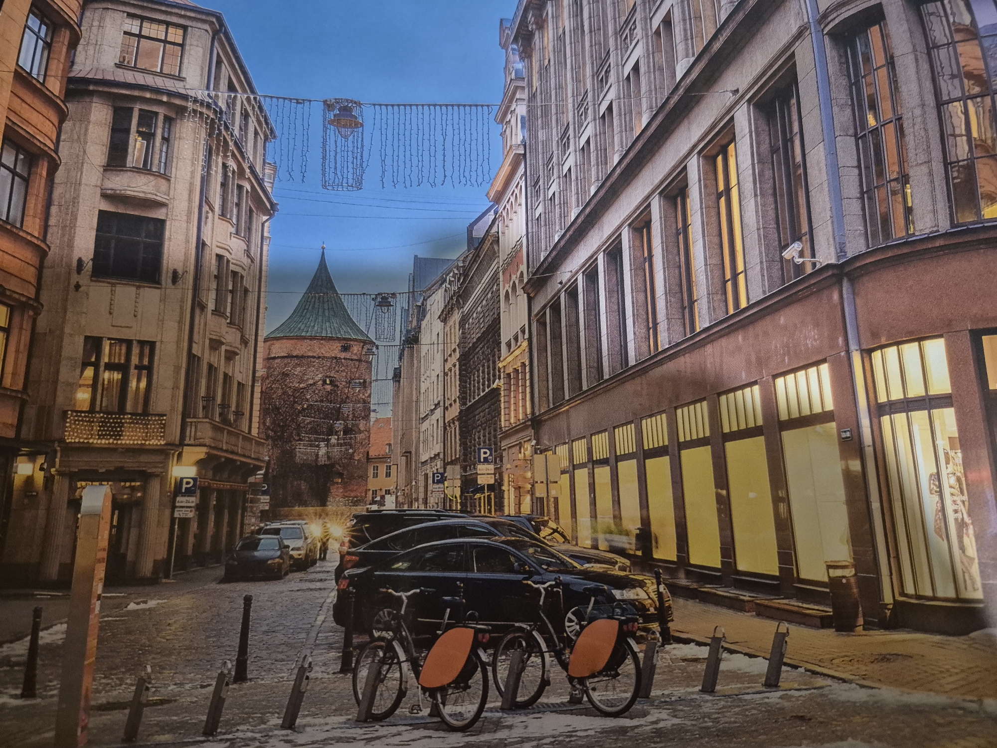 This is a medium shot, eye-level photograph of a cobblestone street in an urban environment during daylight. The street is lined with multi-story buildings, some of which feature classical architecture with large windows and decorative elements. Holiday lights are strung across the street between the buildings, and some of the shops on the ground floor have their lights on. In the foreground, two bicycles are parked, and there are several cars parked along the side of the street. In the background, a tall, conical tower is visible, suggesting a historic district. The sky is a clear, pale blue. There are no people visible in the image.